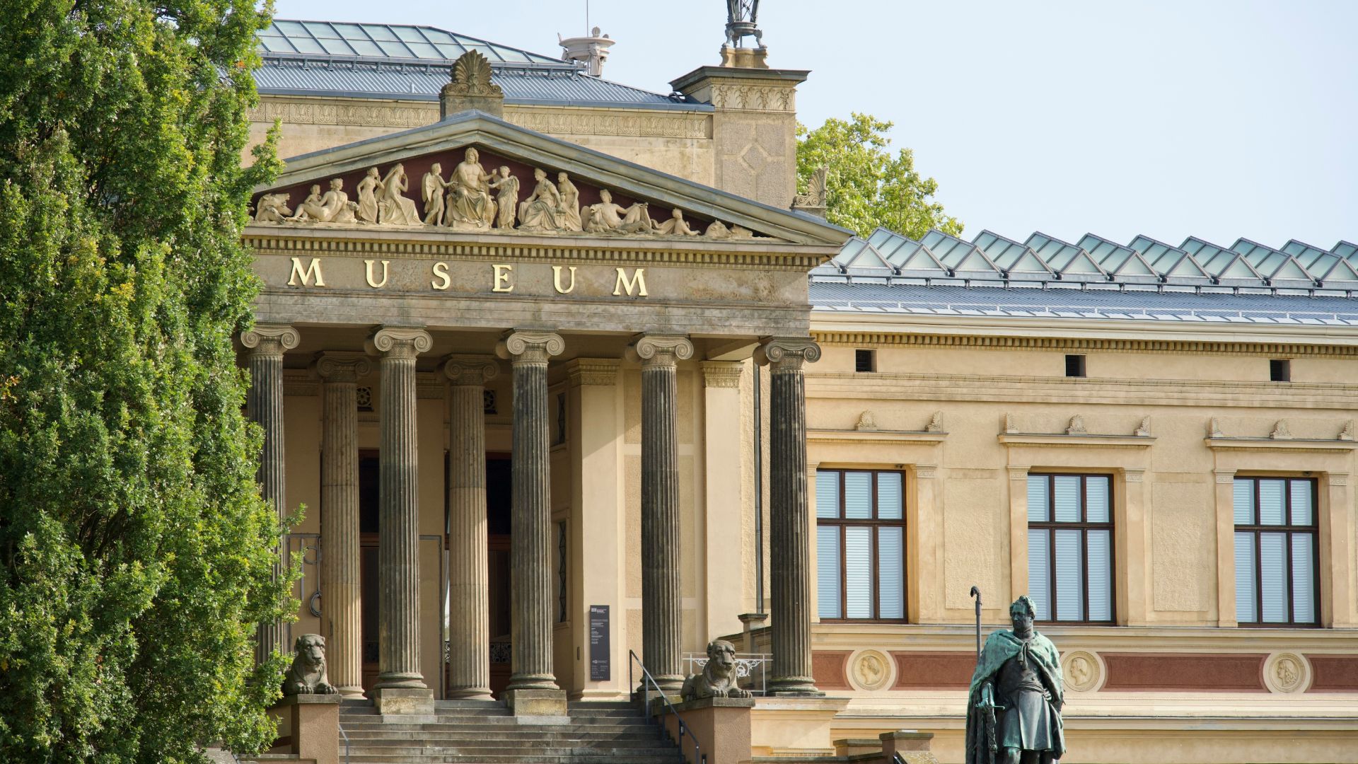 A grand neoclassical museum building with tall columns, a pediment sculpture, a green glass roof, and a statue in the foreground.