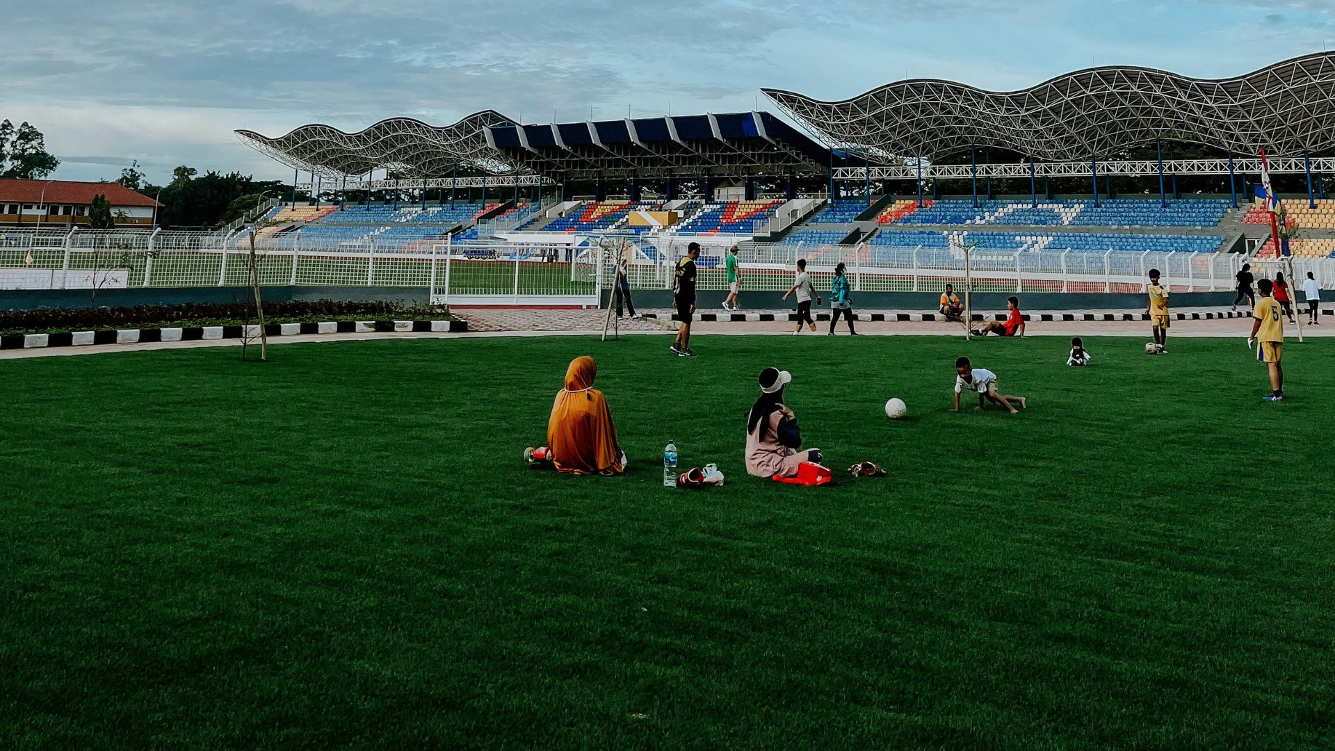 A wide view of stadium field with a modern, blue-seated grandstand and wave-like roof under a bright, cloudy sky, with people relaxing and playing football.