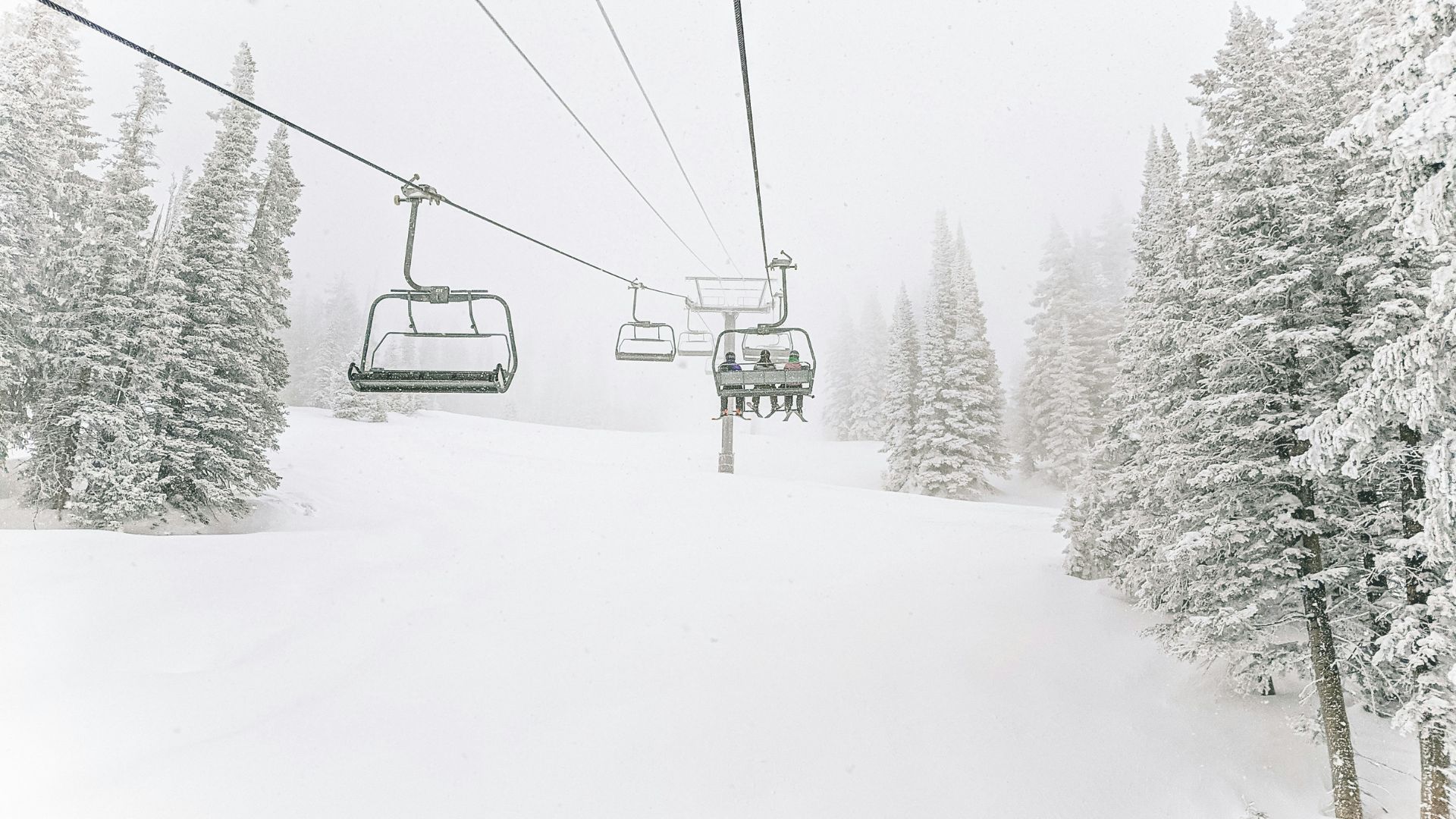 A ski lift carries passengers over a snow-covered slope and past snow-laden evergreen trees during a winter snowstorm at a ski resort.