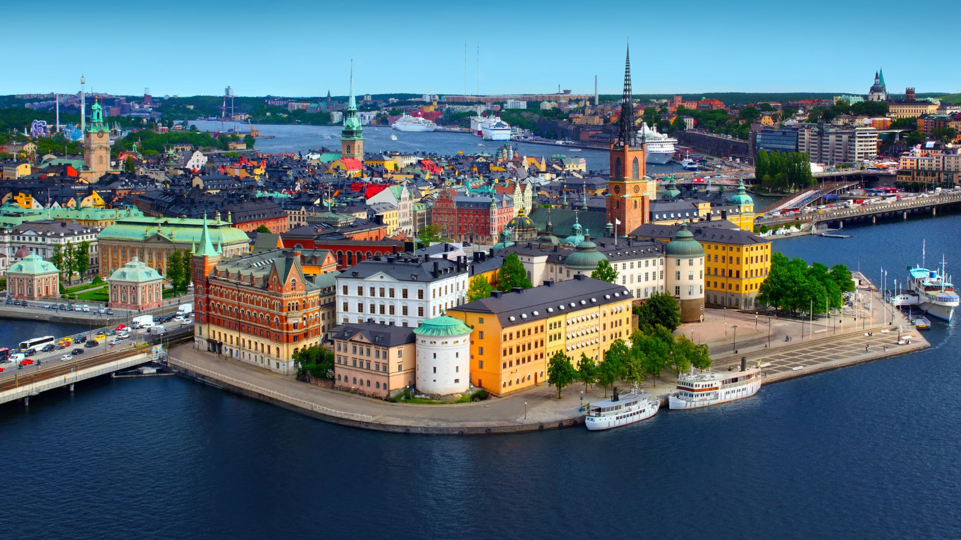 An aerial panorama of the historic Old Town (Gamla Stan) of Stockholm, Sweden, featuring numerous colorful, classic European buildings, several bridges, and church spires towering above the calm blue waters of the harbor on a clear day.