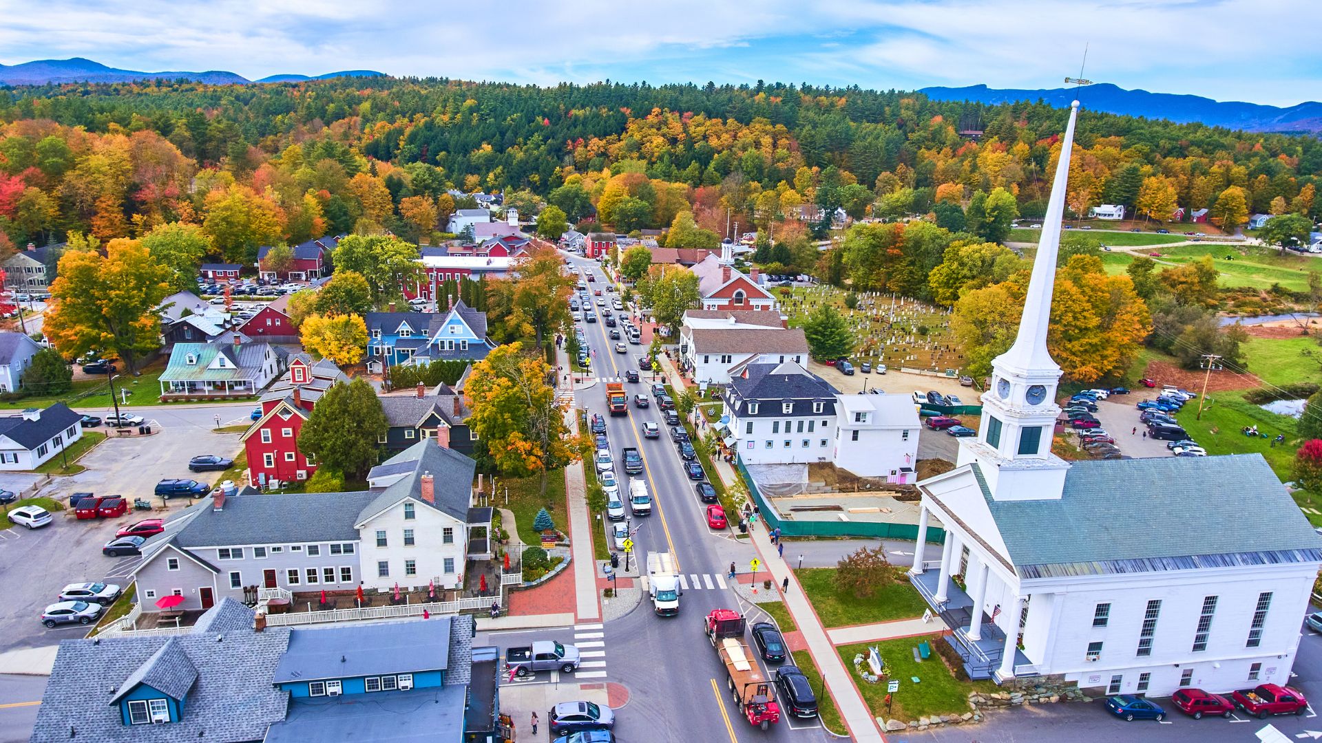 An aerial view of the charming town of Stowe, Vermont, featuring a white church with a tall steeple alongside a main street lined with buildings and autumn trees displaying green, orange, and yellow foliage under a blue sky.