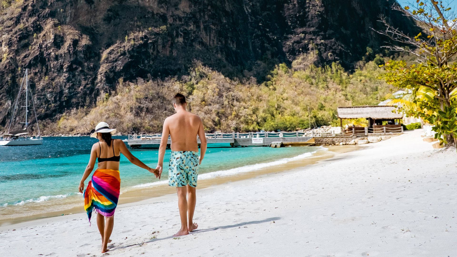 A couple holding hands walks away from the camera along a white sand beach with clear turquoise water and large, lush green mountains in the background.