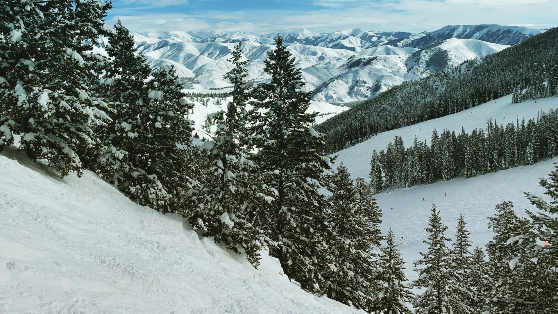 A view from a snowy mountain ski run overlooks a broad valley and multiple layers of distant, snow-covered mountains under a partly cloudy blue sky.