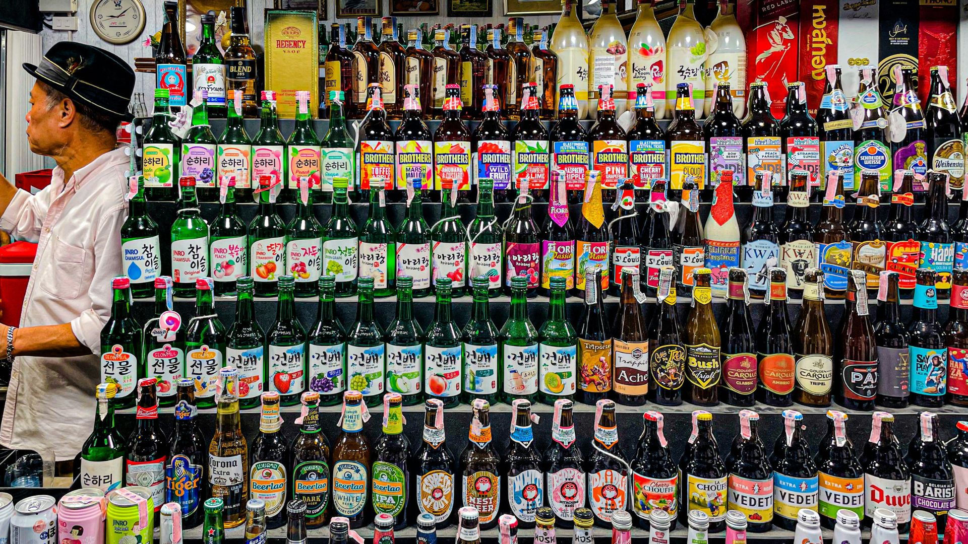 A man stands next to a densely packed, multi-tiered display of various alcohol bottles, including Korean soju brands like 'Sunhari', Thai spirits 'Regency' and 'Hong Thong', and international beers and ciders.
