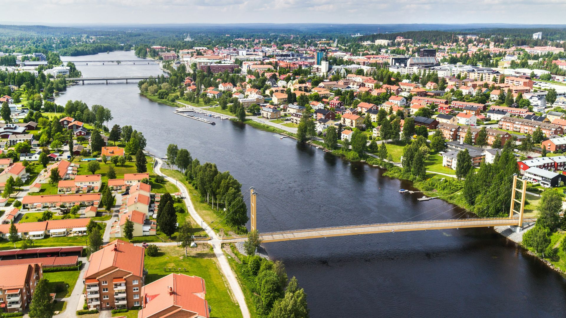 An aerial view of the Swedish city of Skellefteå, showing the dark Skellefte River, a yellow pedestrian bridge, red-roofed buildings, and dense forests under a cloudy sky.