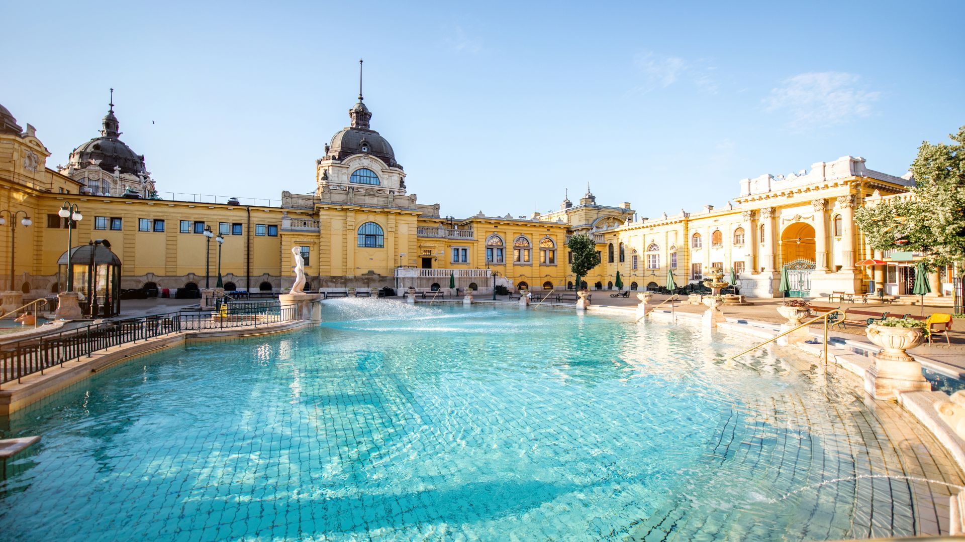 A beautiful, sunny photograph of the grand yellow facade and domes of the historic Széchenyi Thermal Bath in Budapest, Hungary, with its large, turquoise outdoor thermal pool in the foreground.