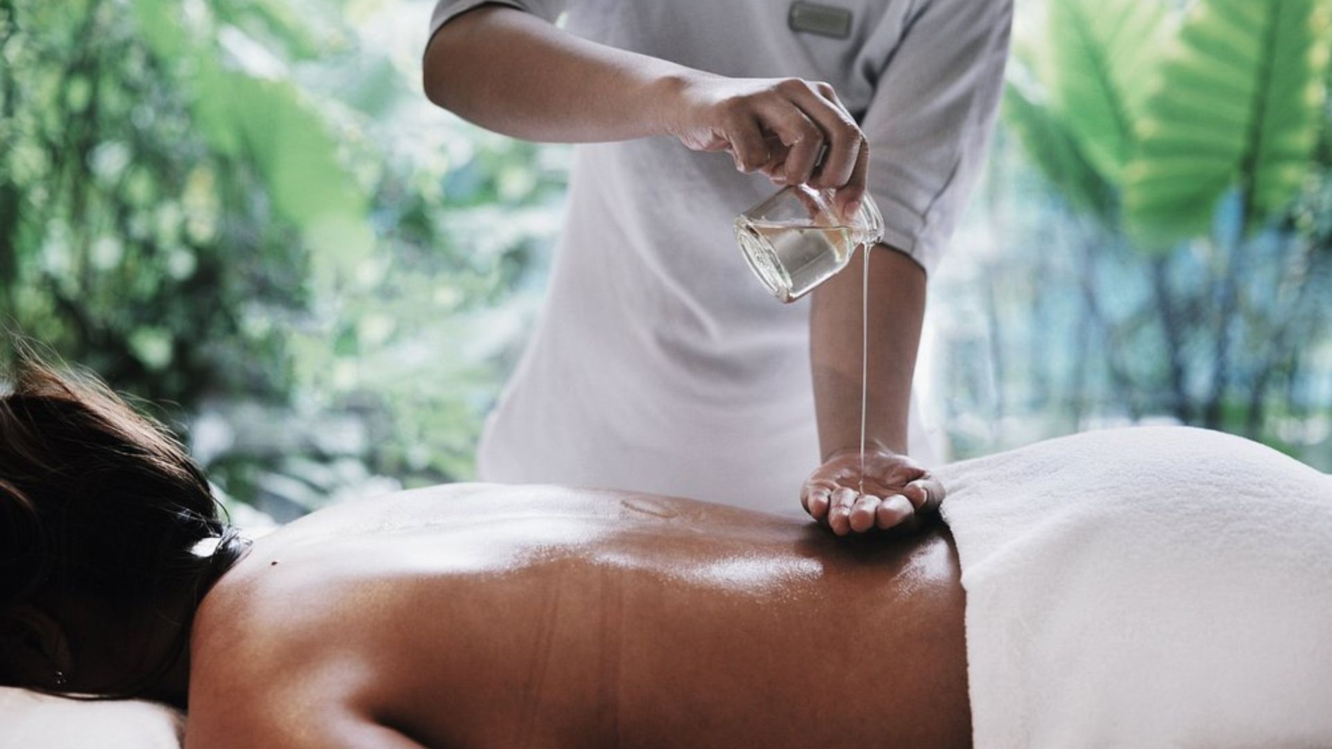 A close-up shot of a spa therapist pouring massage oil onto a person's back during a treatment in a lush, green, outdoor-style setting.