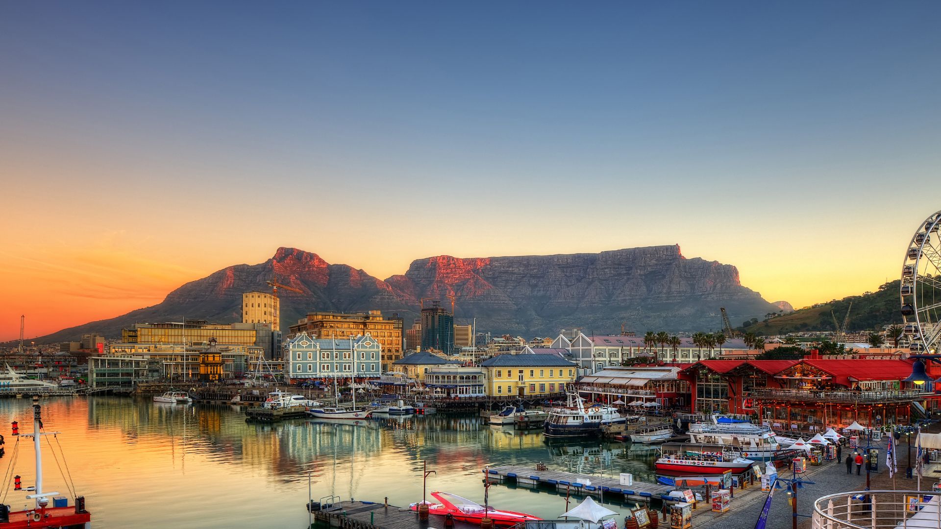 A scenic, sunset view of the V&A Waterfront in Cape Town, South Africa, featuring a bustling harbour, buildings, a ferris wheel, and the iconic Table Mountain in the background.