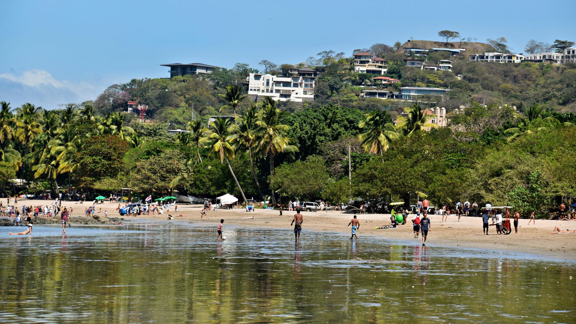 A sunny day at a busy beach with people in the shallow water, palm trees, and houses on a lush green hill in the background.