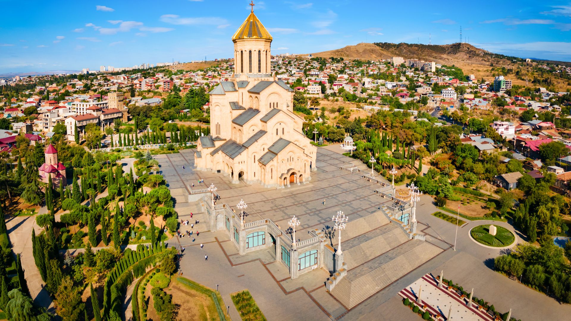 An aerial, panoramic view of the Holy Trinity Cathedral of Tbilisi, Georgia, with its golden dome prominent on a hill overlooking the city.