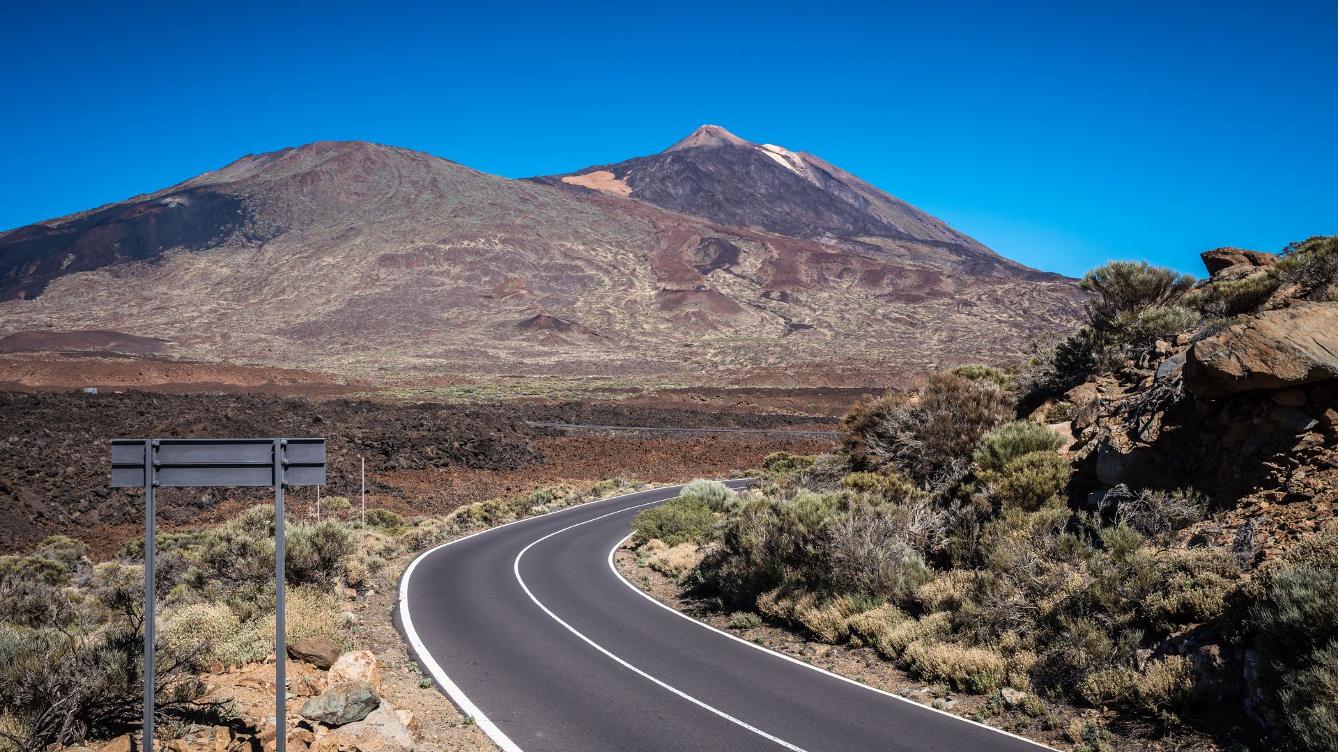 A winding asphalt road cuts through a barren, rocky volcanic landscape, leading toward the imposing Mount Teide volcano under a clear blue sky.