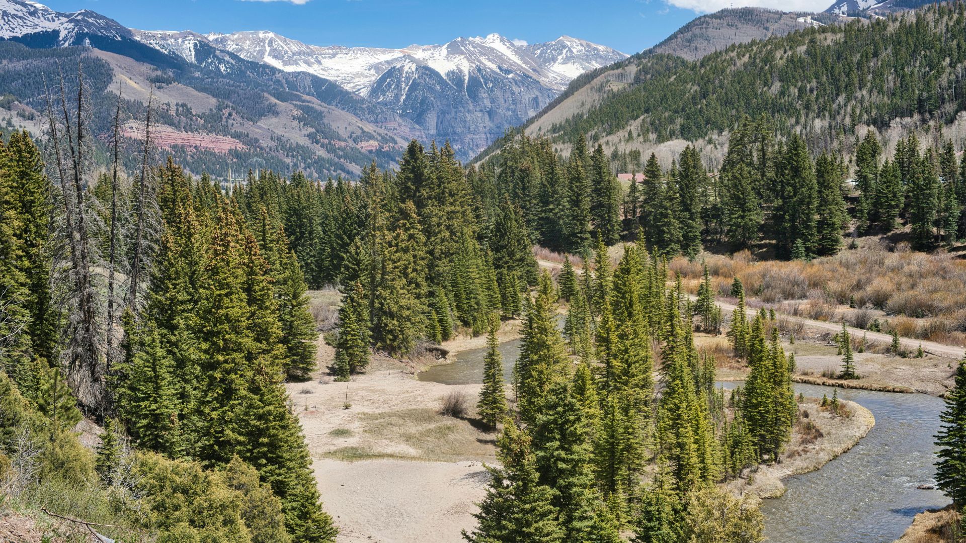 A scenic view of a river winding through a valley filled with evergreen forests, with high, rugged, snow-capped mountains in the background under a blue sky with white clouds.