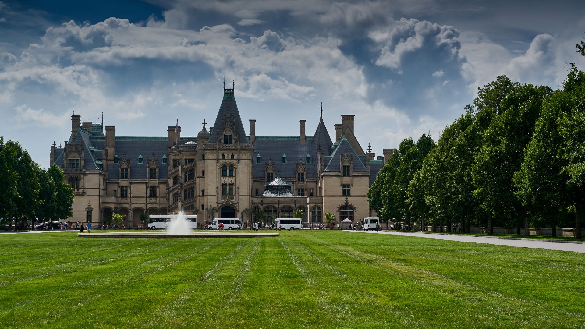 A view of the imposing, French Renaissance-style Biltmore House with its ornate spires and steeply pitched roof, surrounded by expansive green lawns and tall evergreen trees under a blue sky with white clouds.