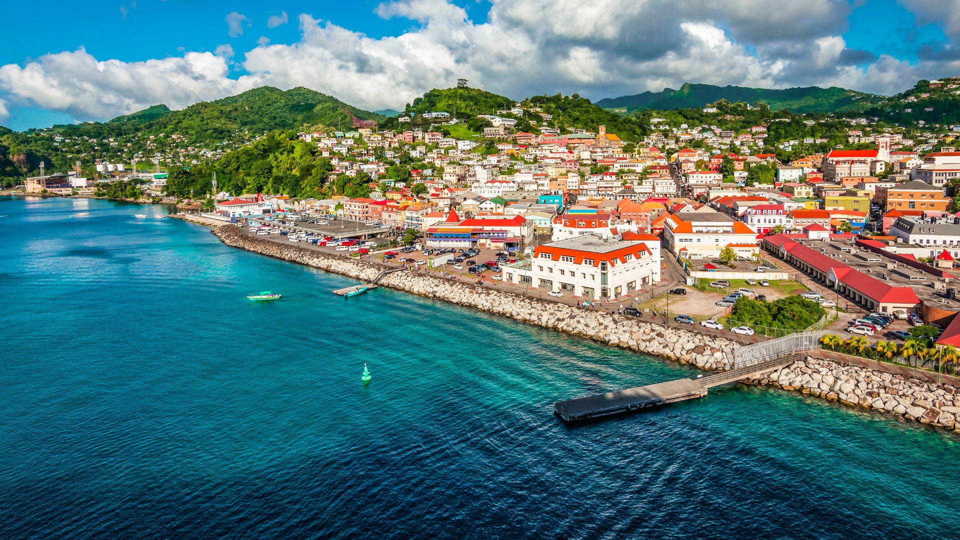 A bright, elevated view of The Carenage waterfront in St. George's, Grenada, showing a harbor with turquoise water, a long stone quay, and a hillside town with colorful buildings.
