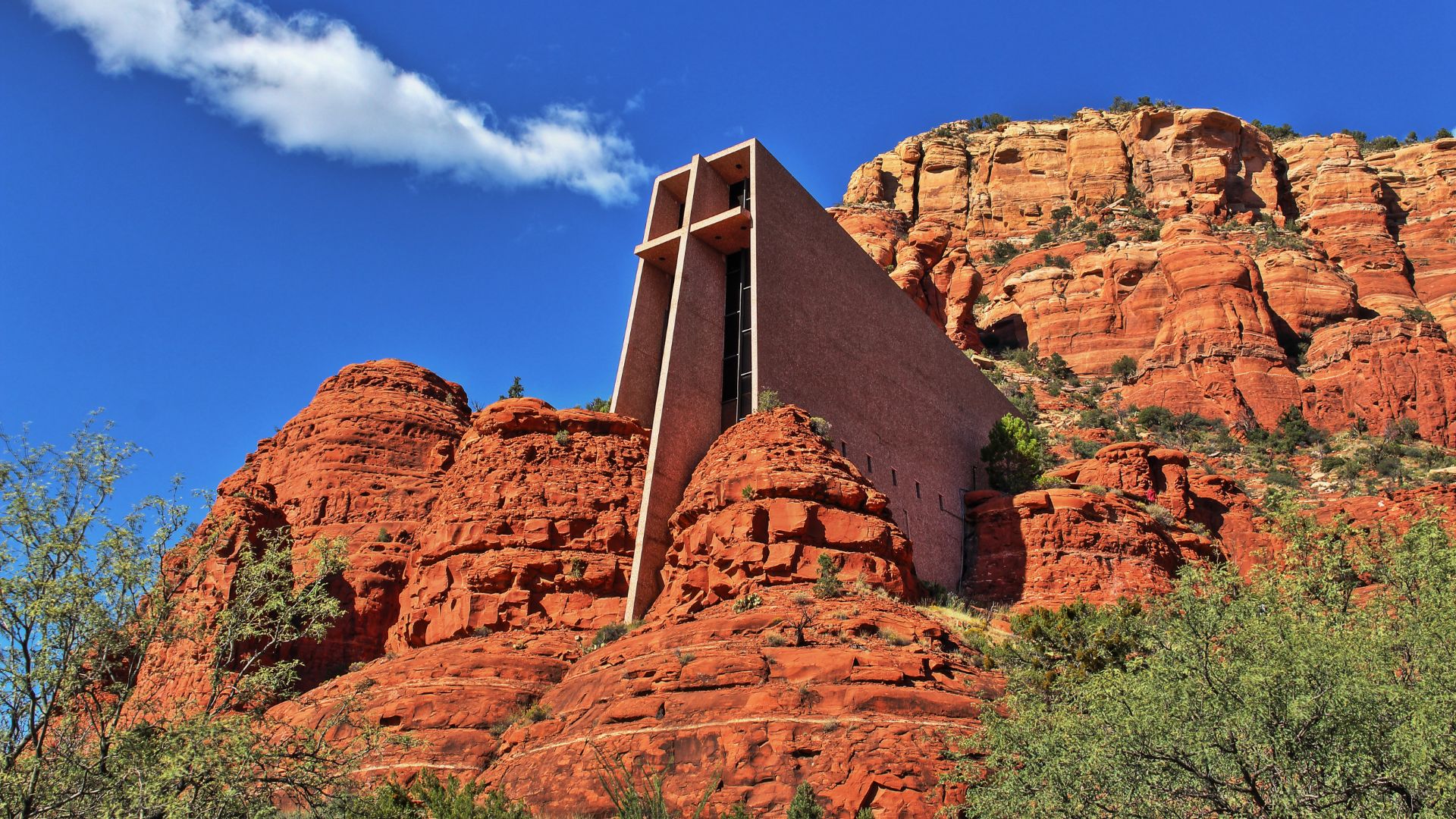 A striking modernist chapel with a large cross integrated into its design is built directly into towering red rock buttes under a blue sky with white clouds in Sedona, Arizona.