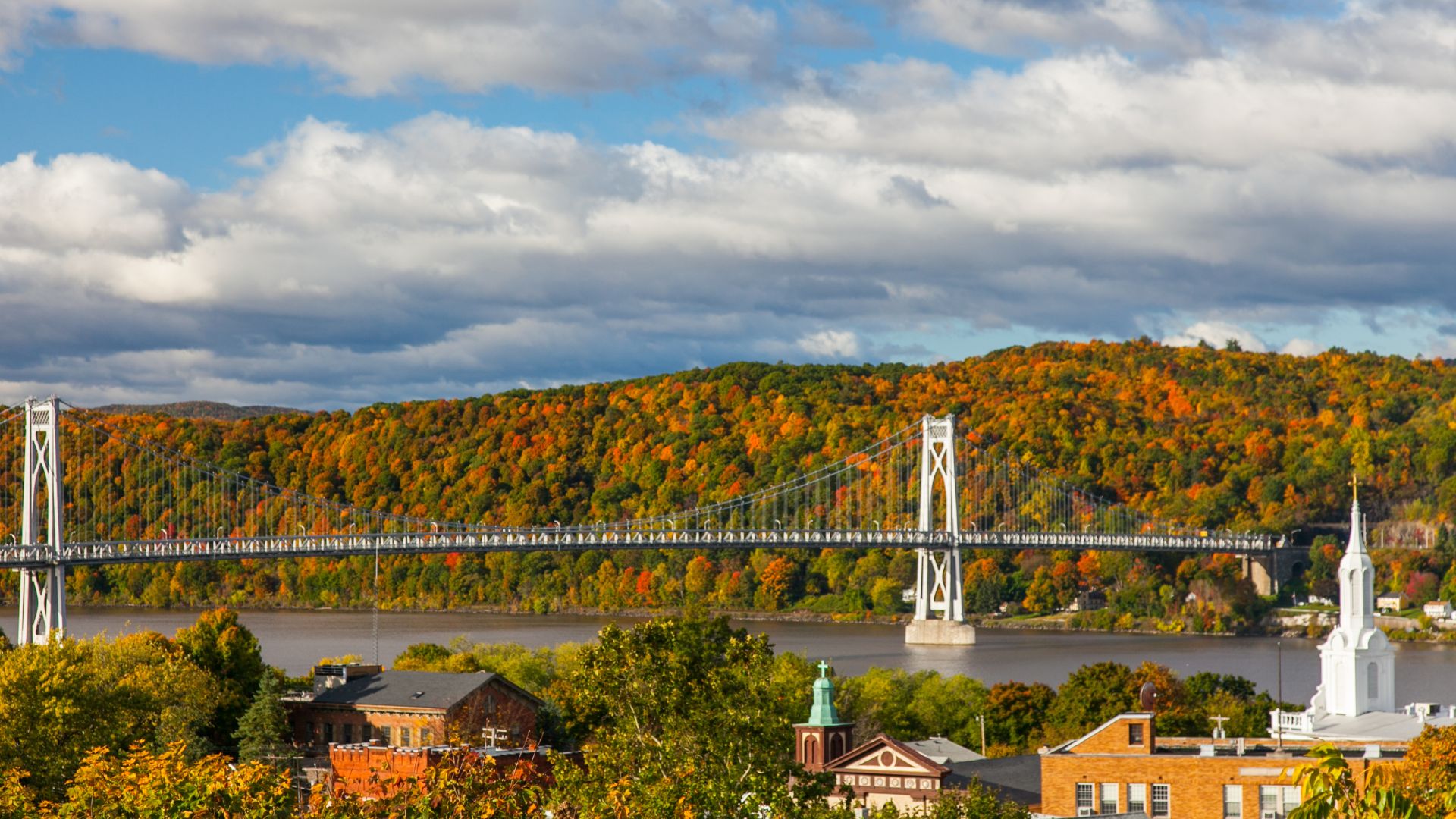 A suspension bridge over a wide river, with a town in the foreground and hills covered in vibrant autumn foliage under a partly cloudy sky.