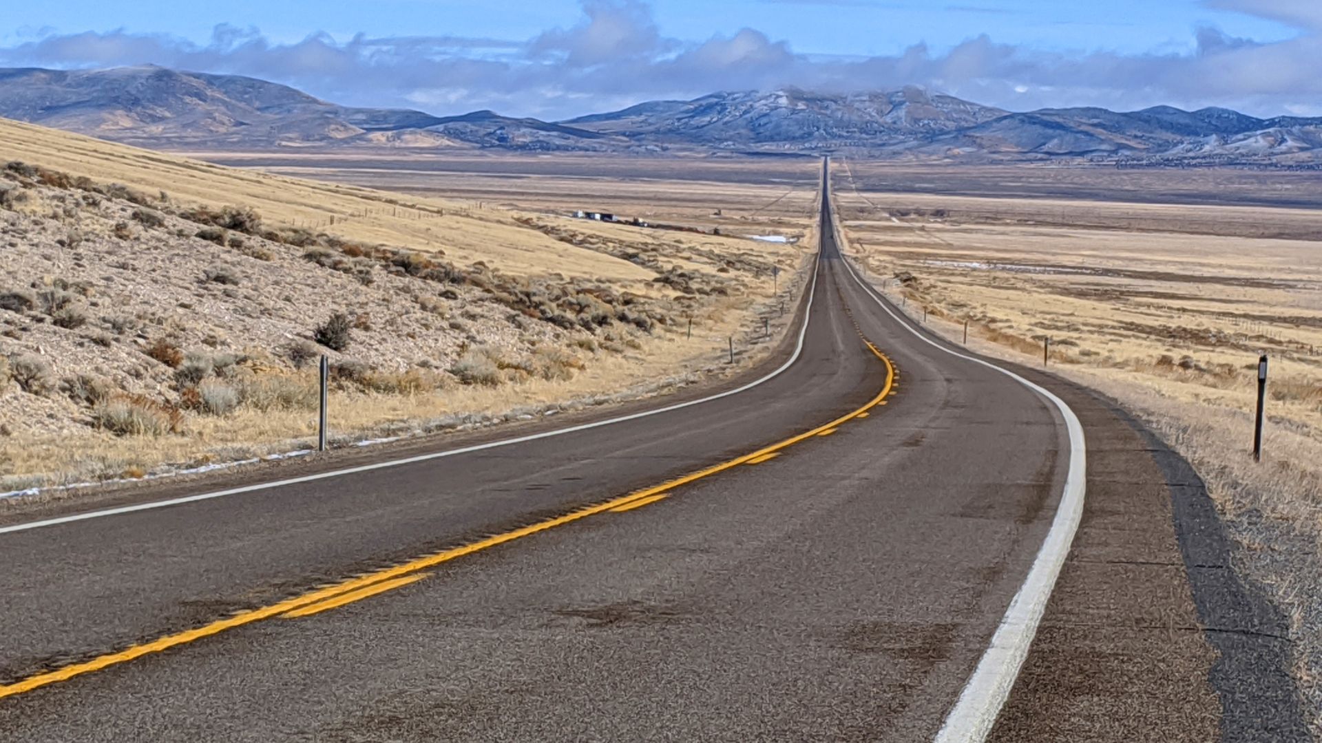 A two-lane paved highway curves into the distance through a wide, arid desert valley with dry, golden hillsides and distant snow-capped mountains under a bright blue sky with scattered white clouds.