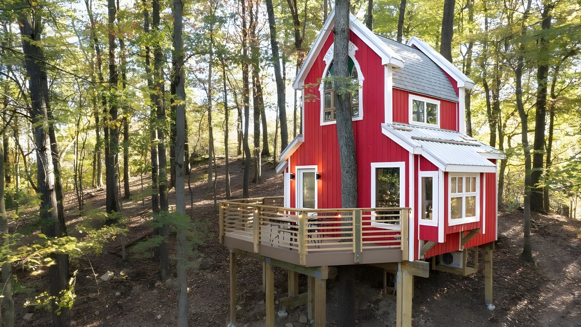 A bright red, two-story treehouse with white trim and a wrap-around wooden deck is built high into a forested, autumnal hillside.