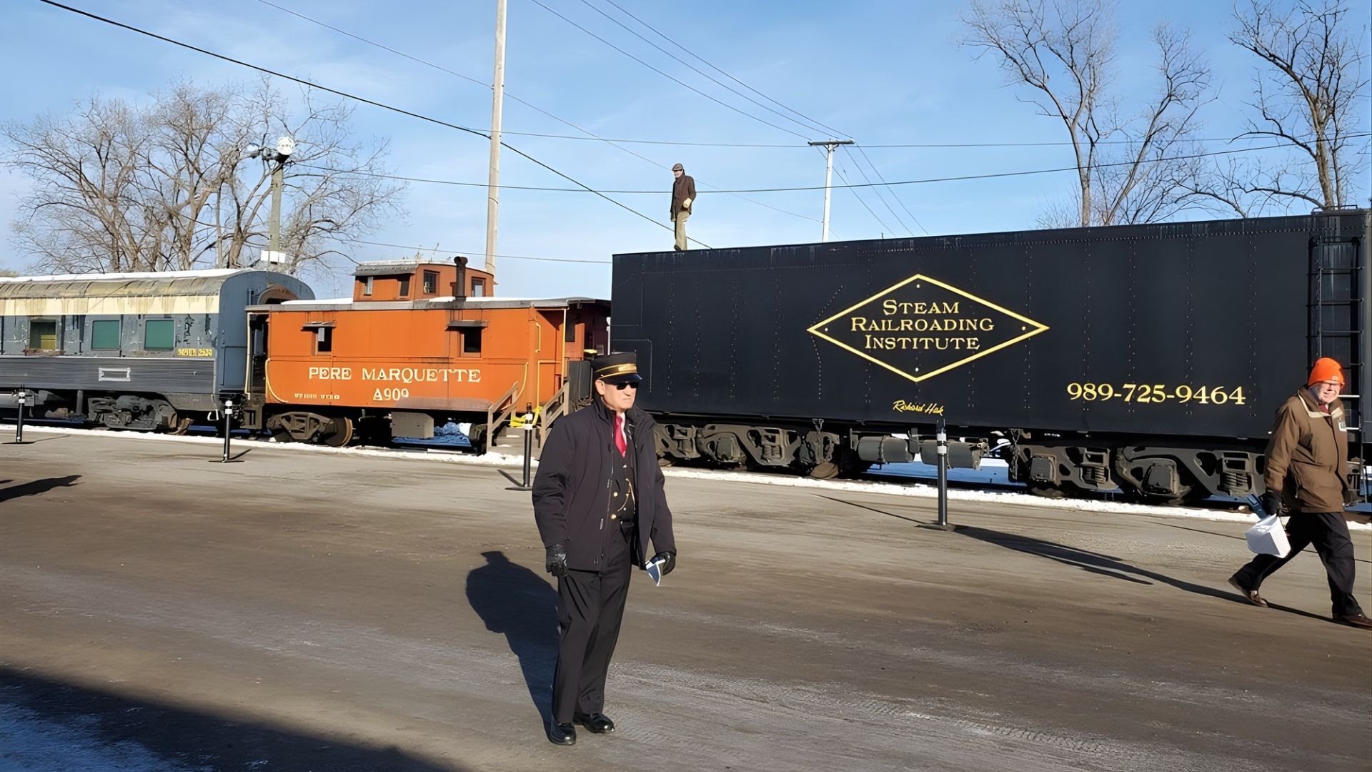 A train yard scene at the Steam Railroading Institute in Owosso, Michigan, featuring several vintage rail cars including the Pere Marquette A900, and people in the foreground.