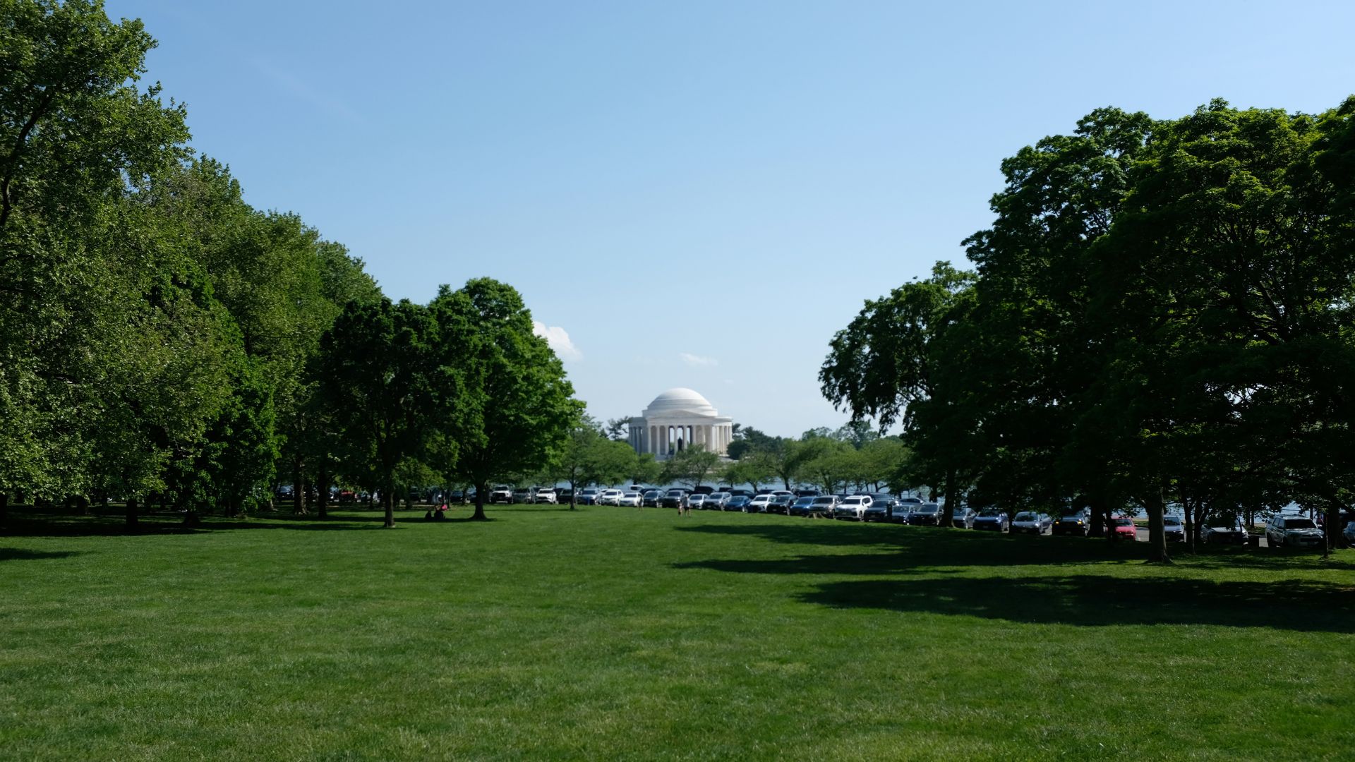 A wide shot across a green park lawn towards the domed Thomas Jefferson Memorial building in the distance, with a line of parked cars visible among the trees along the water's edge under a clear blue sky.