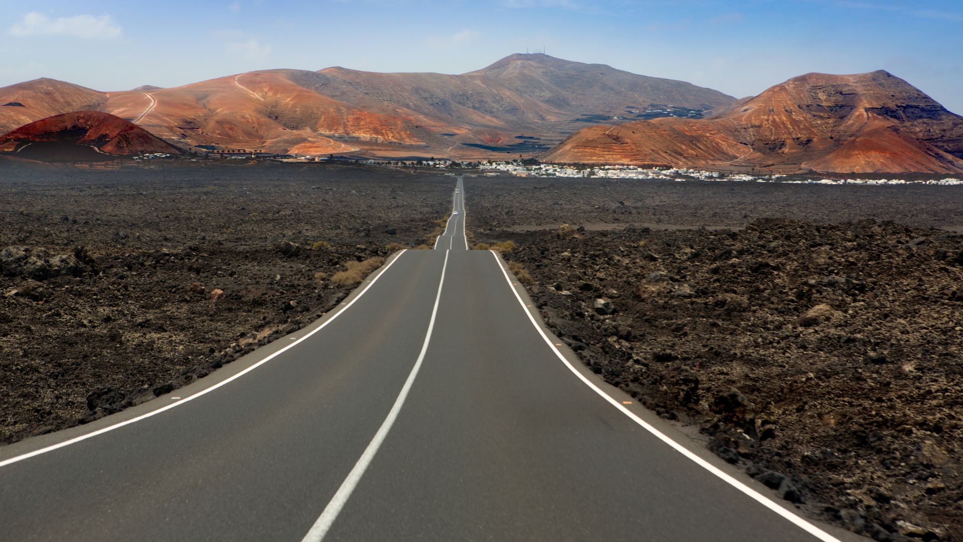 A straight road cuts through a vast, dark volcanic landscape towards reddish-brown mountains under a clear sky