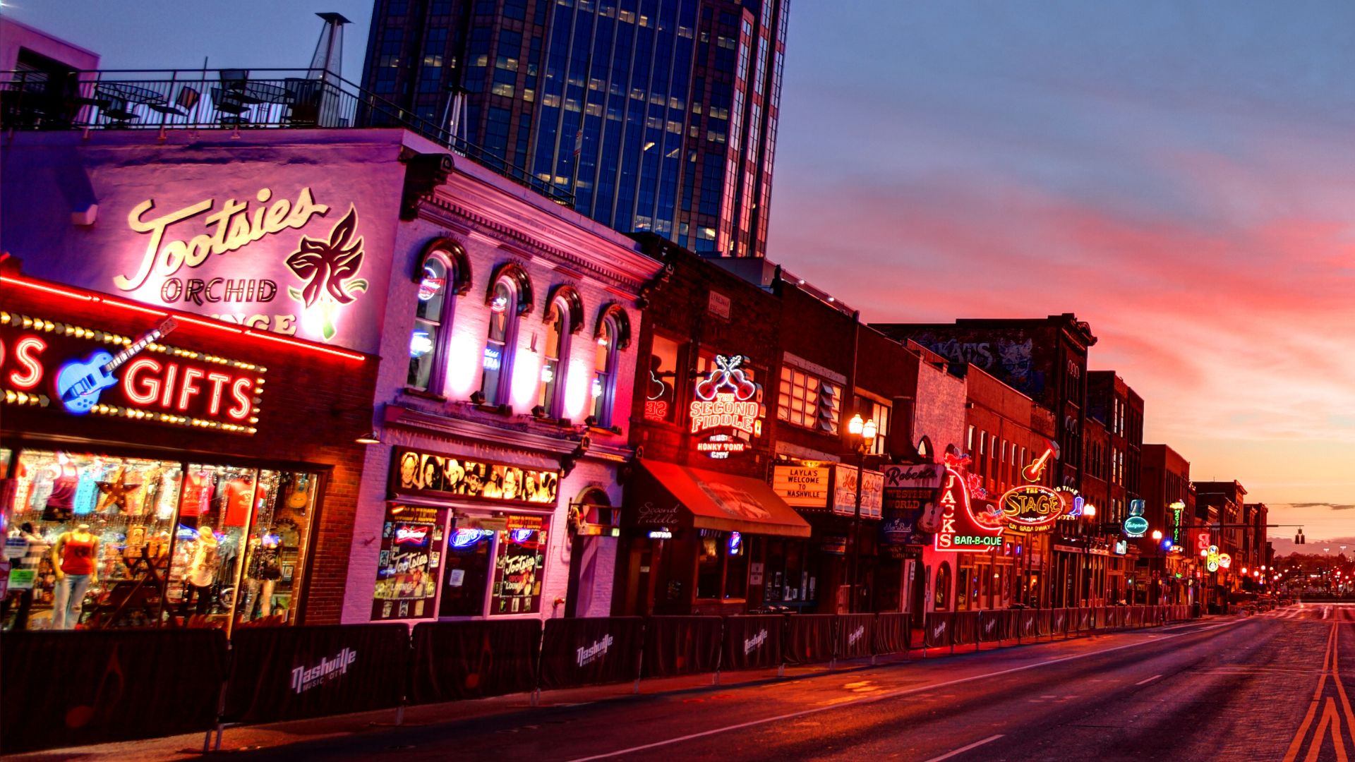 A photograph taken at sunset showing the brightly lit neon signs of several honky-tonk bars along Broadway street in Downtown Nashville, Tennessee, with the most prominent sign reading "Tootsies Orchid Lounge"