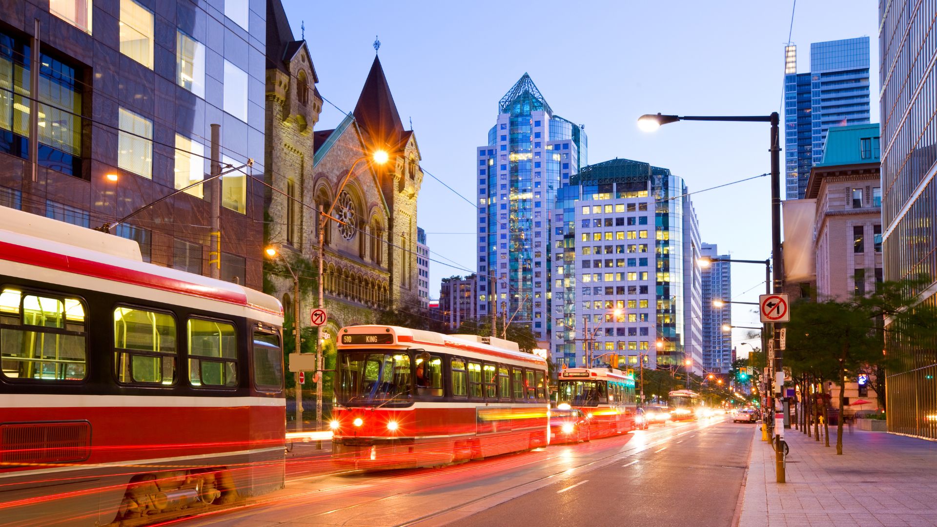 A night-time street scene in downtown Toronto, Canada, showing two red TTC streetcars with illuminated headlights on a street lined with modern skyscrapers and a historic church building.