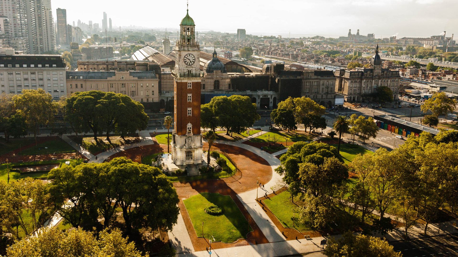 A large, brick clock tower with a green dome and a clock face dominates a city park with winding paths and trees, surrounded by dense city buildings.