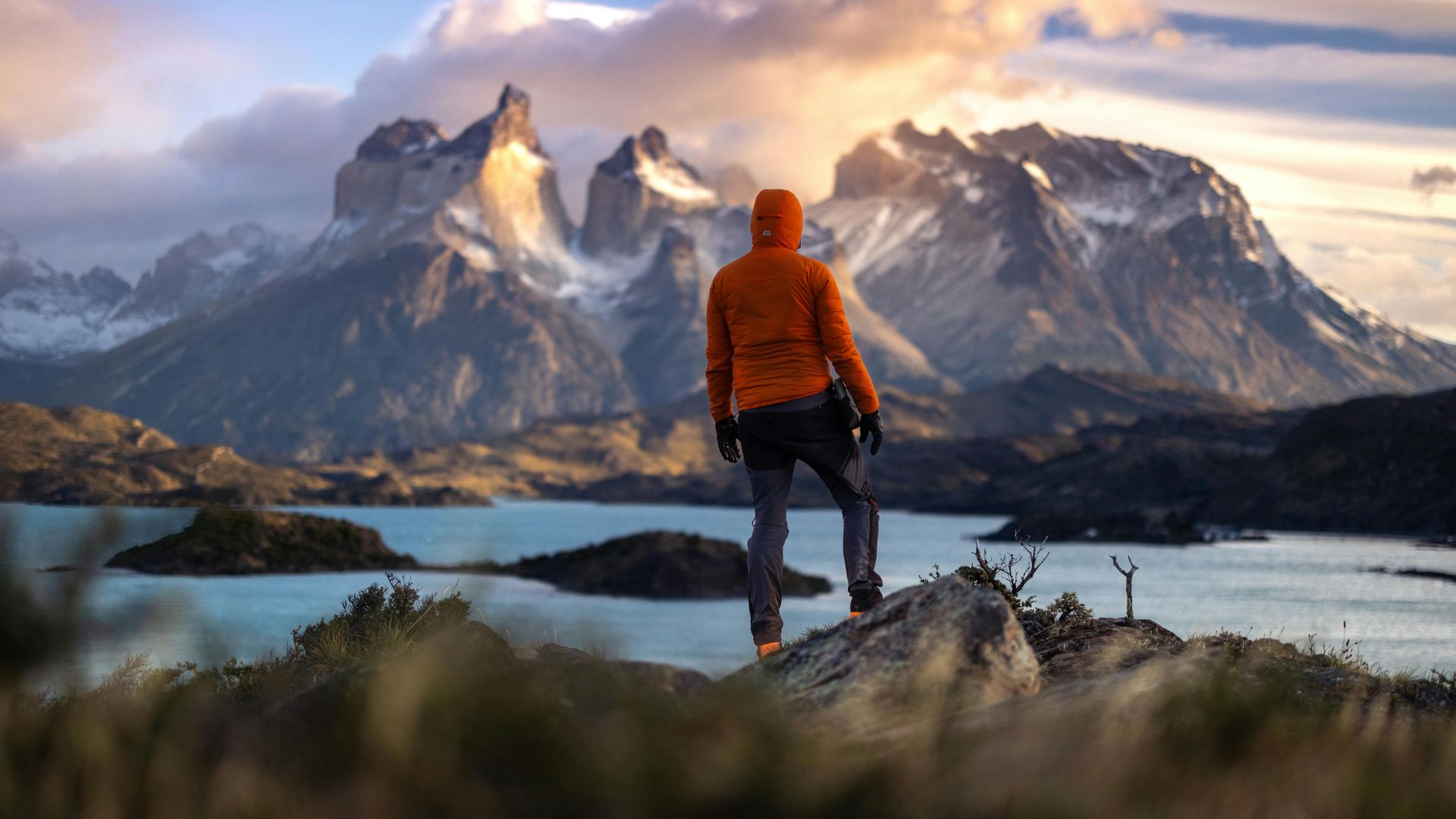 A person wearing an orange jacket stands on a rock overlooking a lake and the majestic, snow-capped granite peaks of Torres del Paine National Park at sunset.