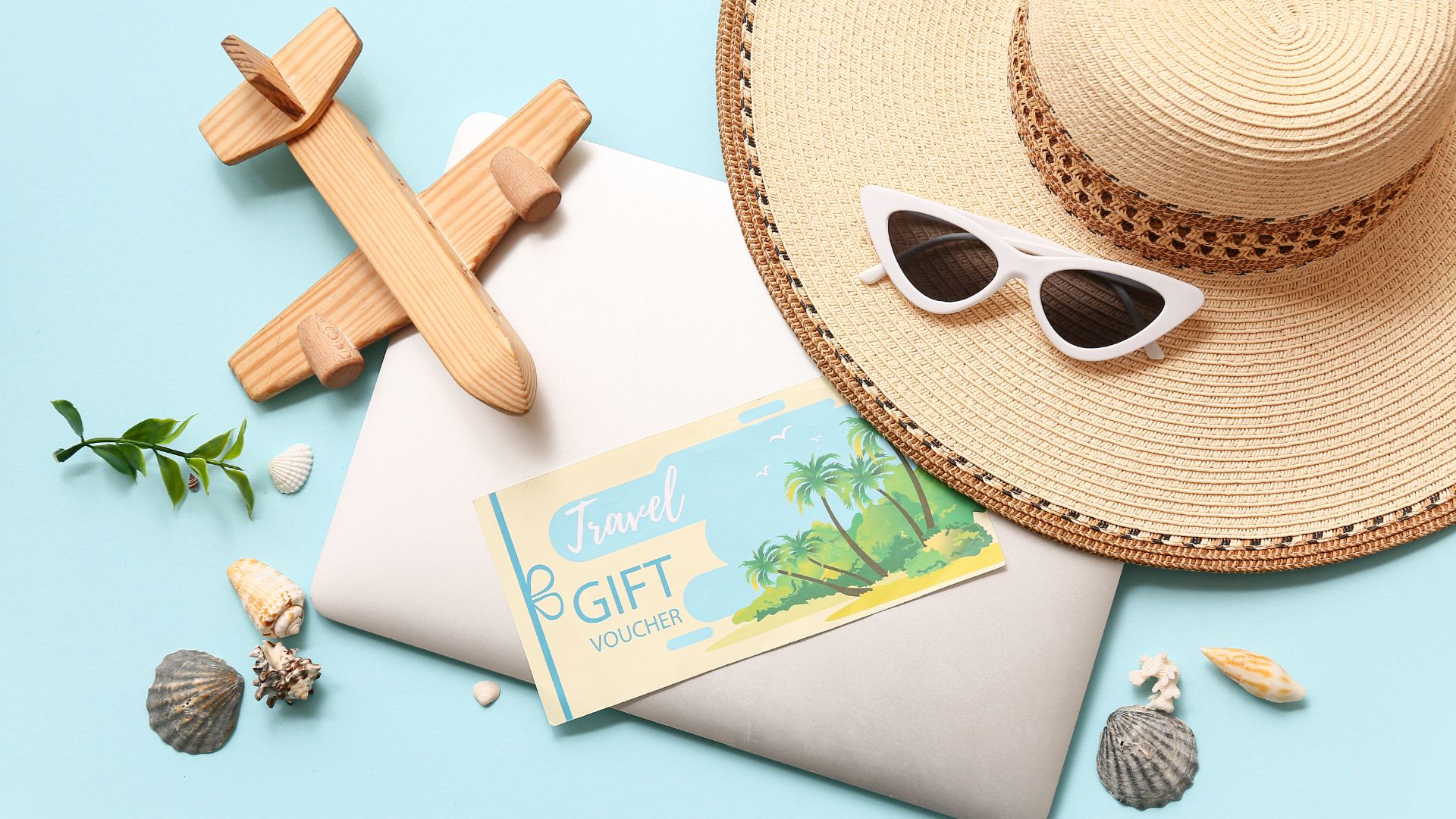 A flat lay arrangement of travel items including a wooden toy airplane, a sun hat, white cat-eye sunglasses, a travel gift voucher, and assorted seashells on a light blue background.