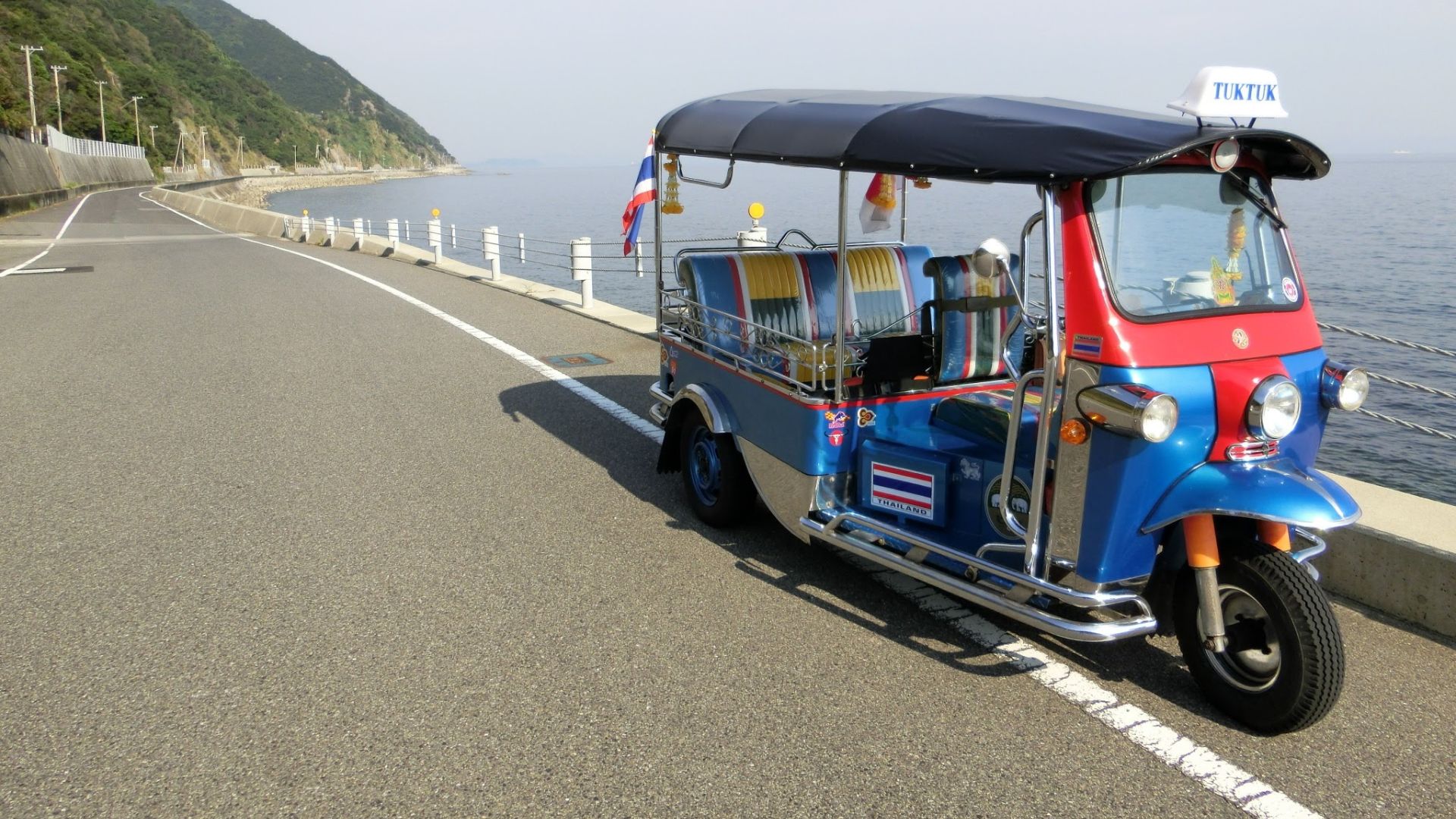 A colorful Thai-style tuk-tuk parked on a coastal road overlooking the sea.