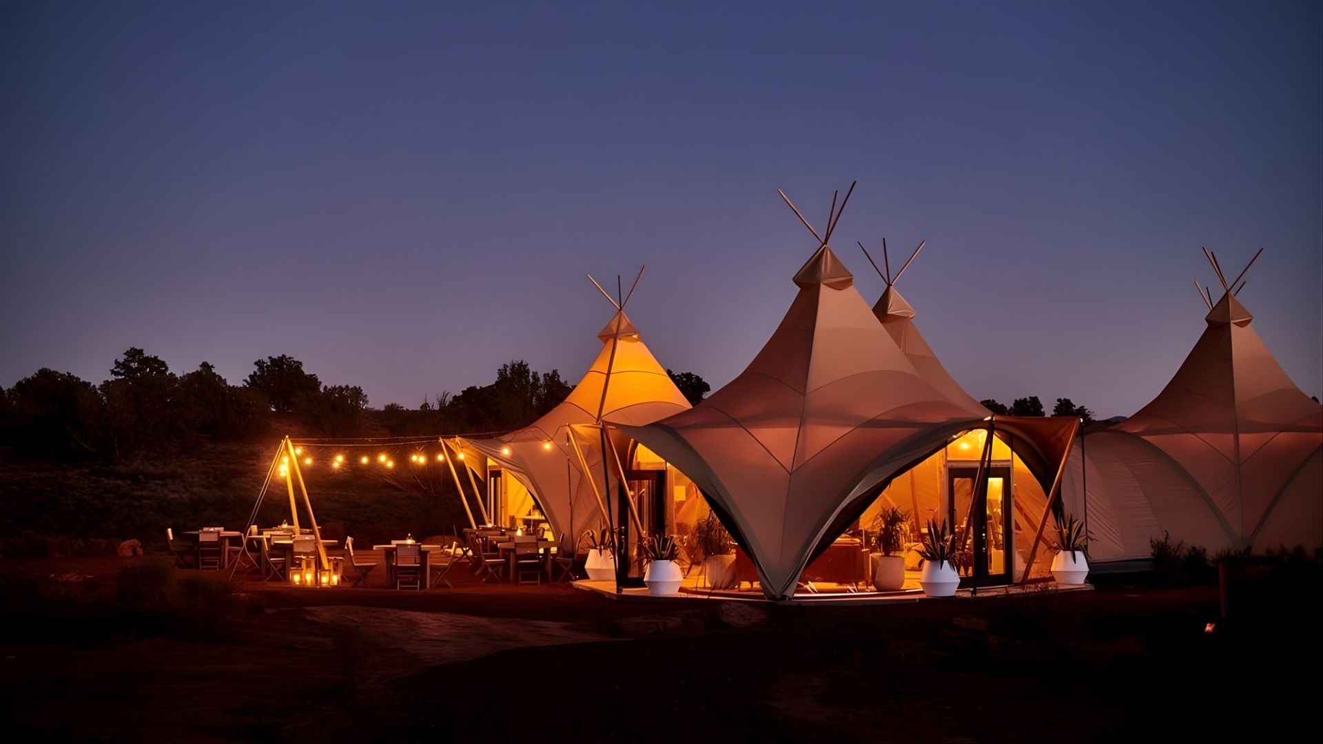 A nighttime photo of an outdoor glamping resort featuring several large, illuminated canvas tents and an outdoor dining area with string lights under a dark sky.