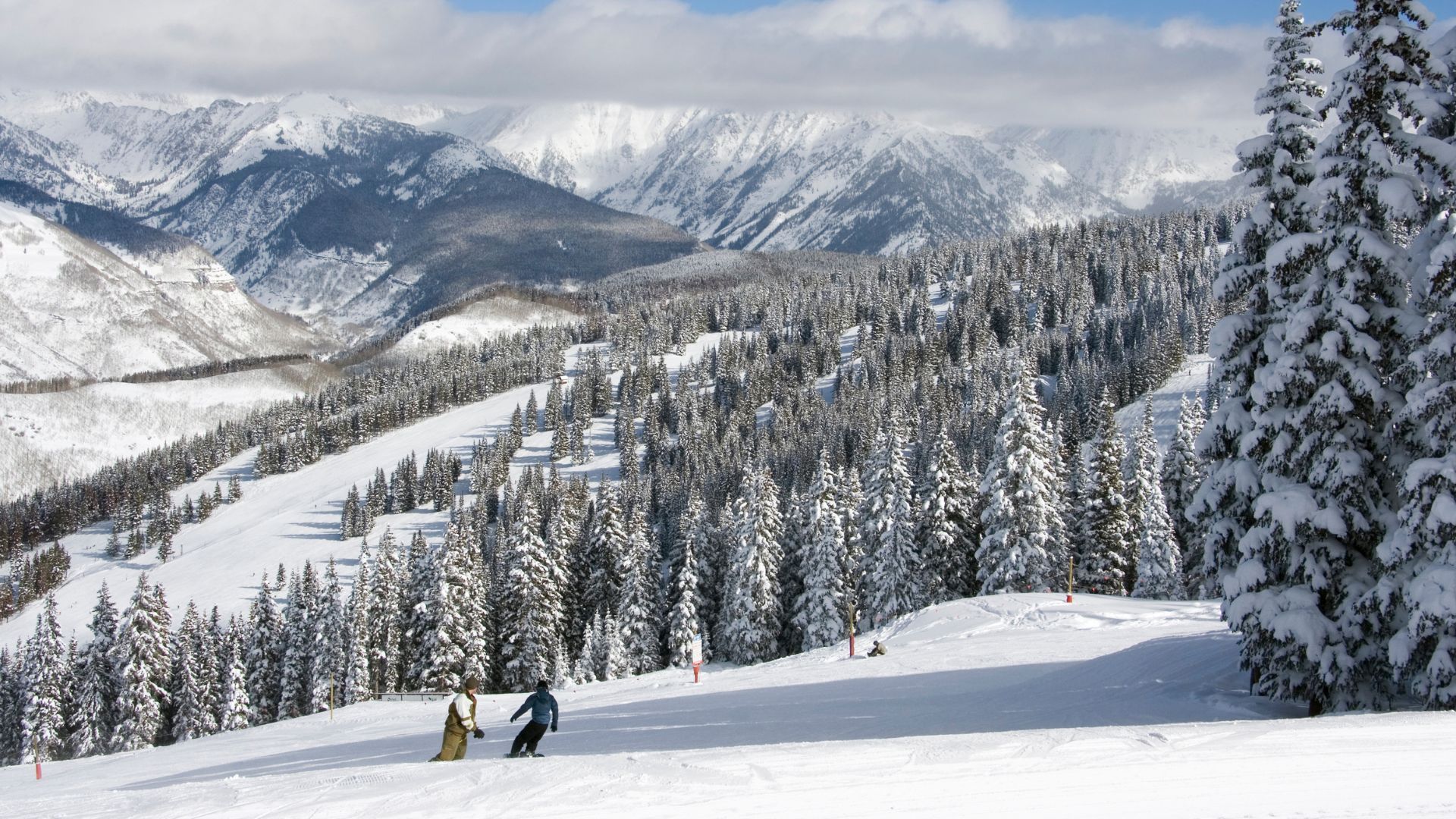 A view from a wide, groomed ski slope looking across a vast evergreen forest toward a jagged, snow-capped mountain range in Vail, Colorado.