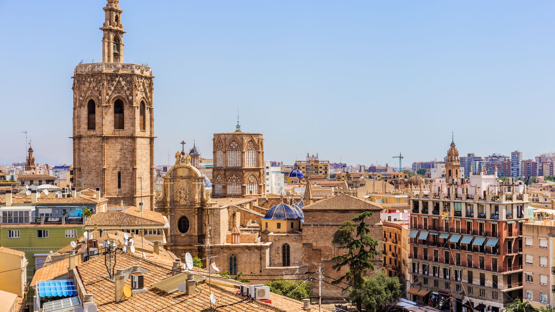 An elevated, daytime photograph of the historic rooftops of Valencia, Spain, with the ornate, octagonal Miguelete bell tower of the Valencia Cathedral rising prominently on the left under a clear blue sky.