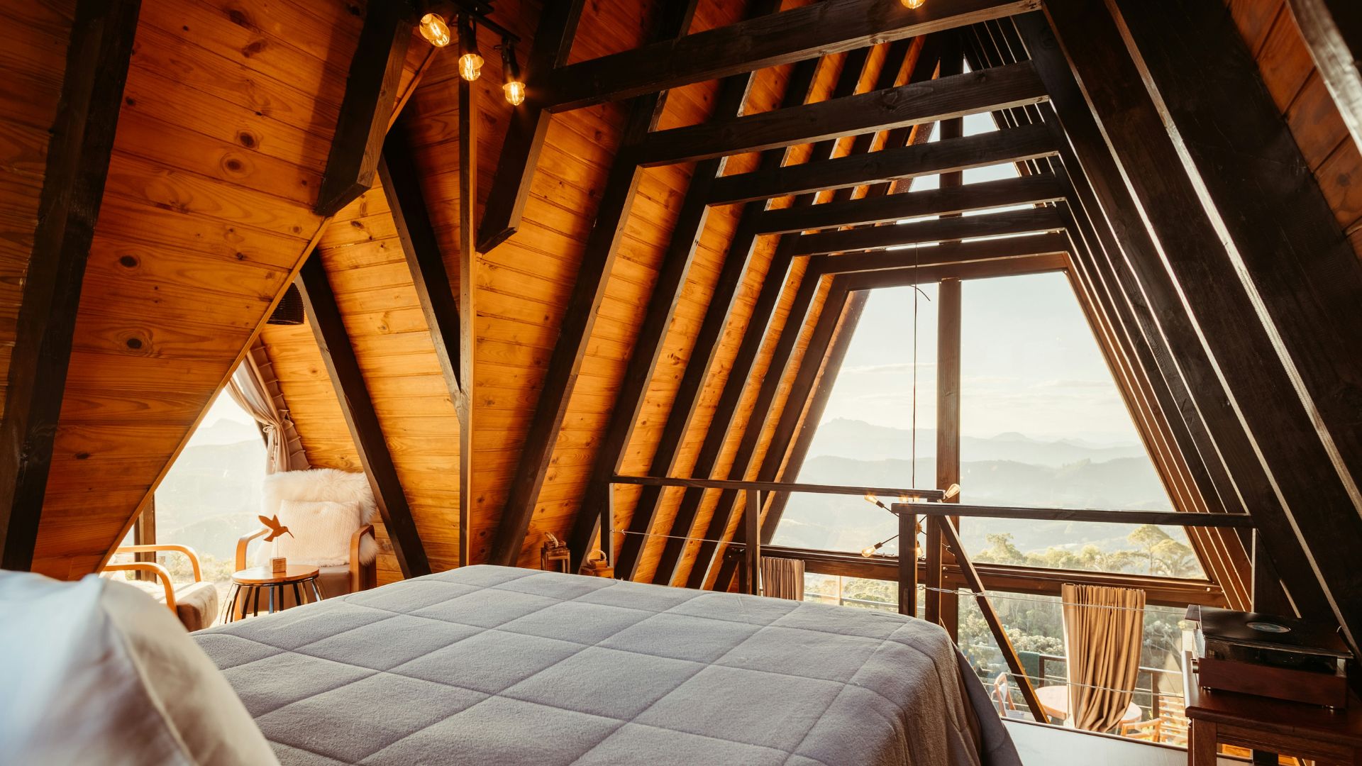 The interior of a rustic A-frame cabin bedroom with a large bed, exposed wooden beams, and a wide window view of a mountain landscape.