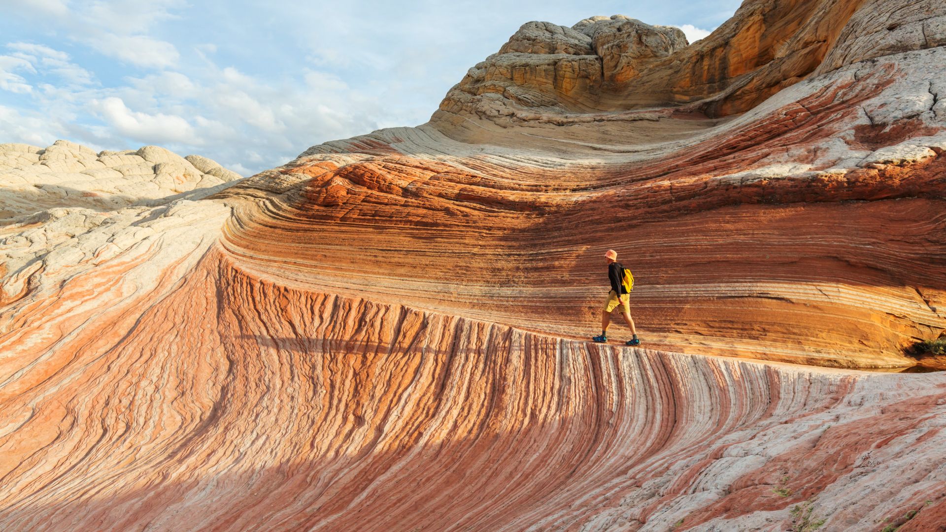 A man with a backpack hikes along a massive, naturally formed ridge of swirling red and white sandstone rock formations under a bright sky.