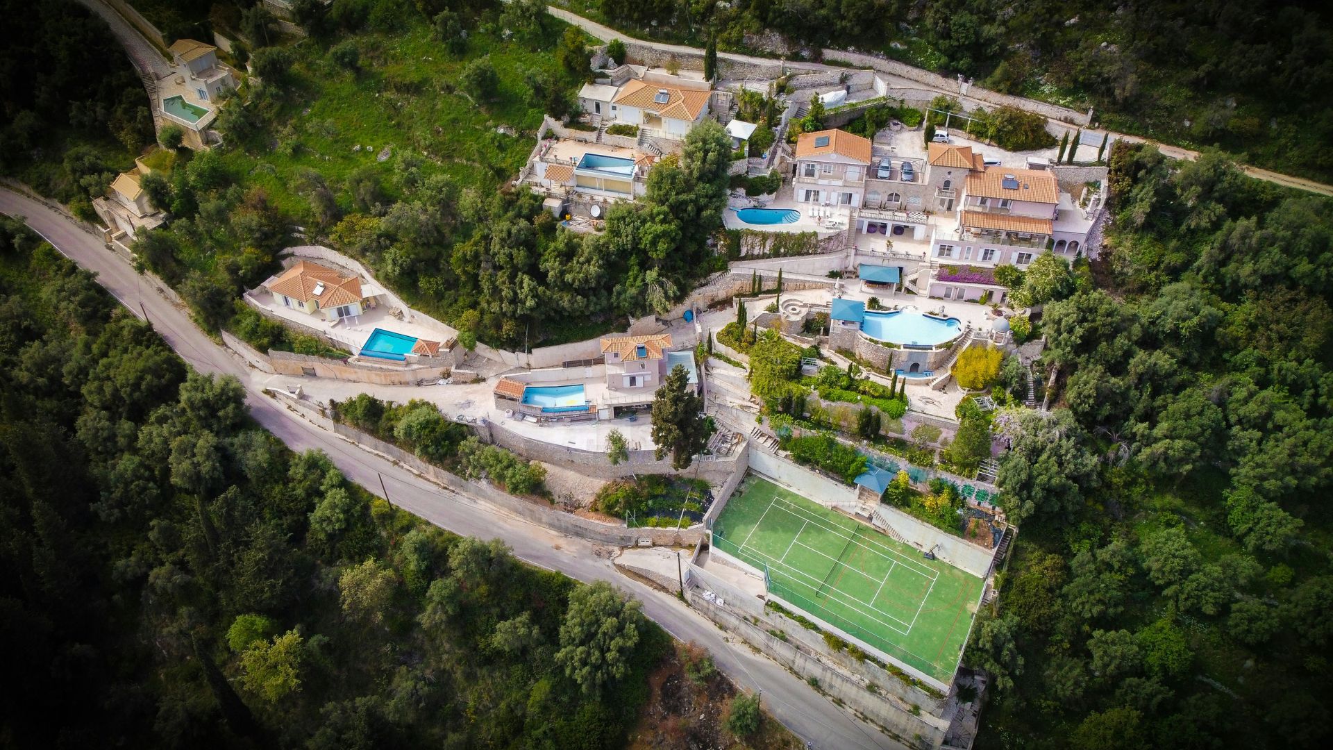 An aerial view of a luxury villa complex with multiple swimming pools and a tennis court nestled on a steep, tree-covered hillside.
