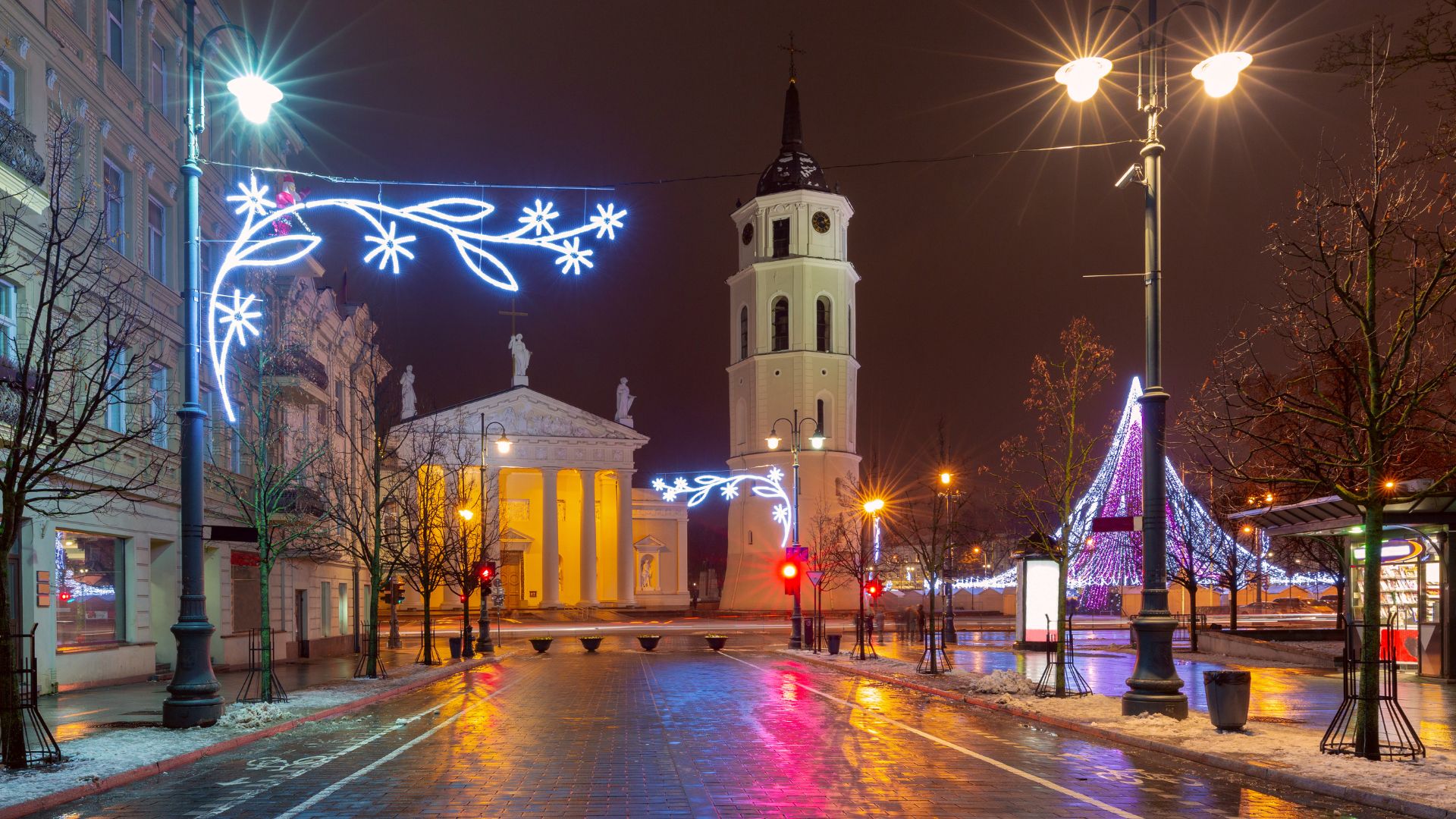 A night view of the white, classicist Vilnius Cathedral and its tall, detached bell tower, illuminated by warm lights and festive blue and white Christmas decorations on a wet street.