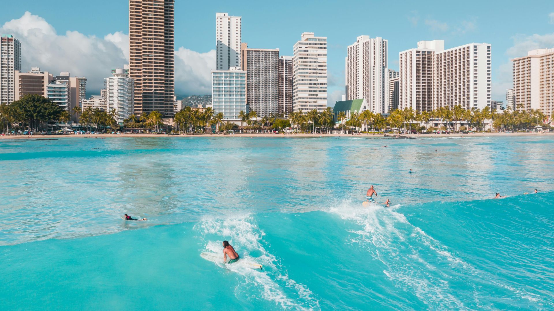 Aerial view of people surfing on turquoise waves at Waikiki Beach, with a skyline of high-rise hotels and buildings in the background.