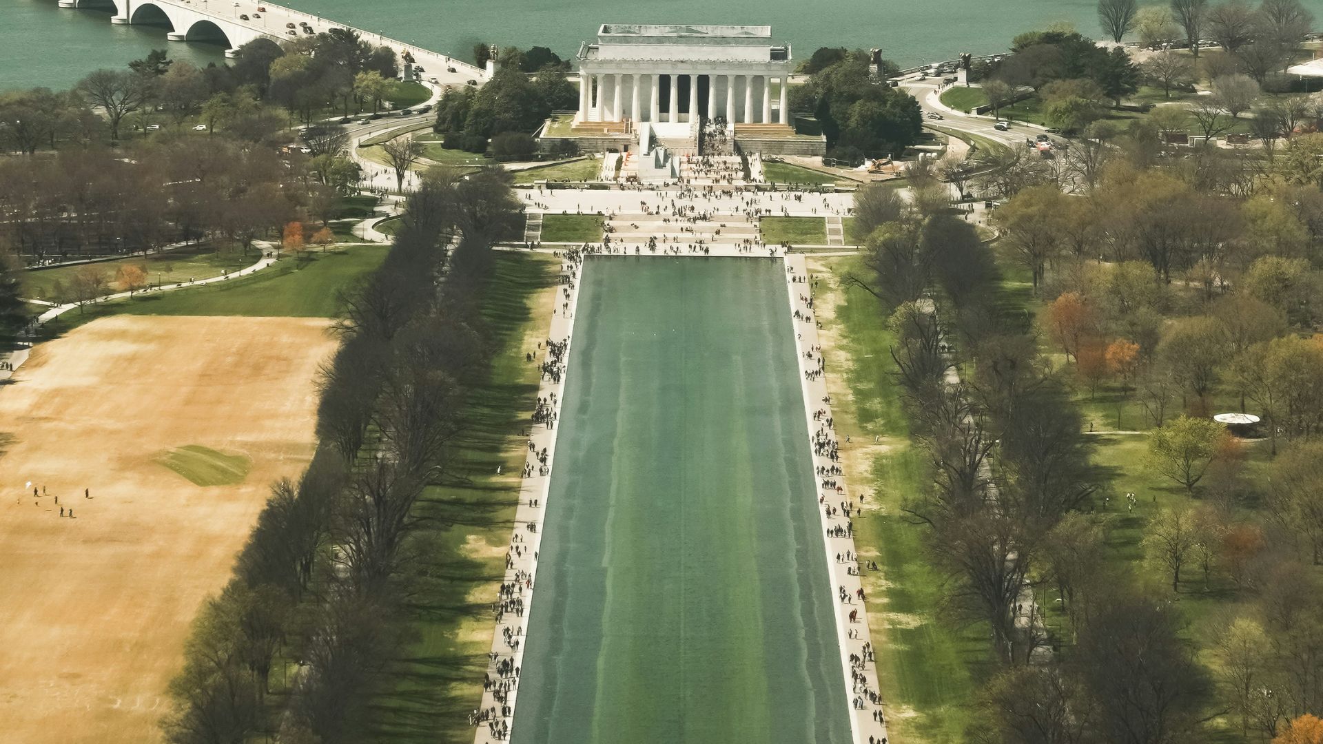 An aerial view looking west down the long Lincoln Memorial Reflecting Pool toward the Lincoln Memorial, with the white Arlington Memorial Bridge spanning the Potomac River in the background.