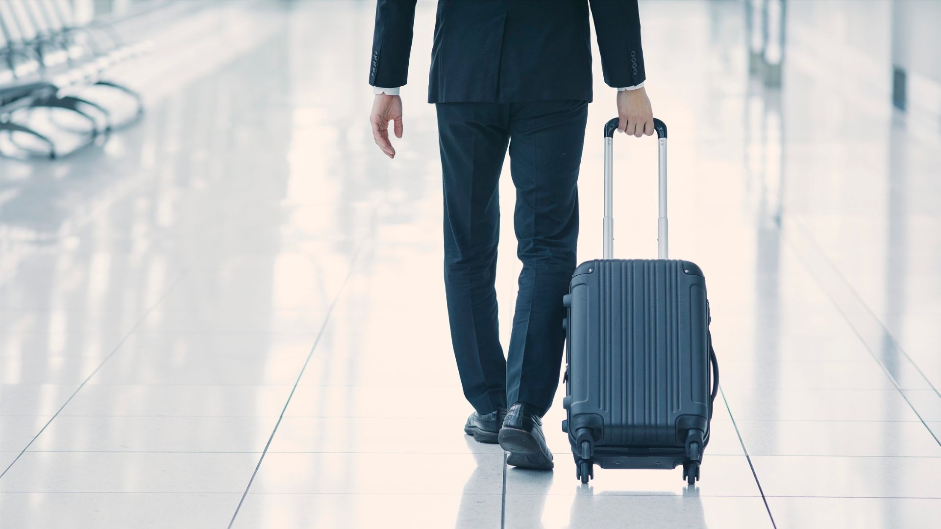 A person in a suit is walking away down a bright airport terminal while pulling a blue-gray hardside rolling carry-on suitcase.