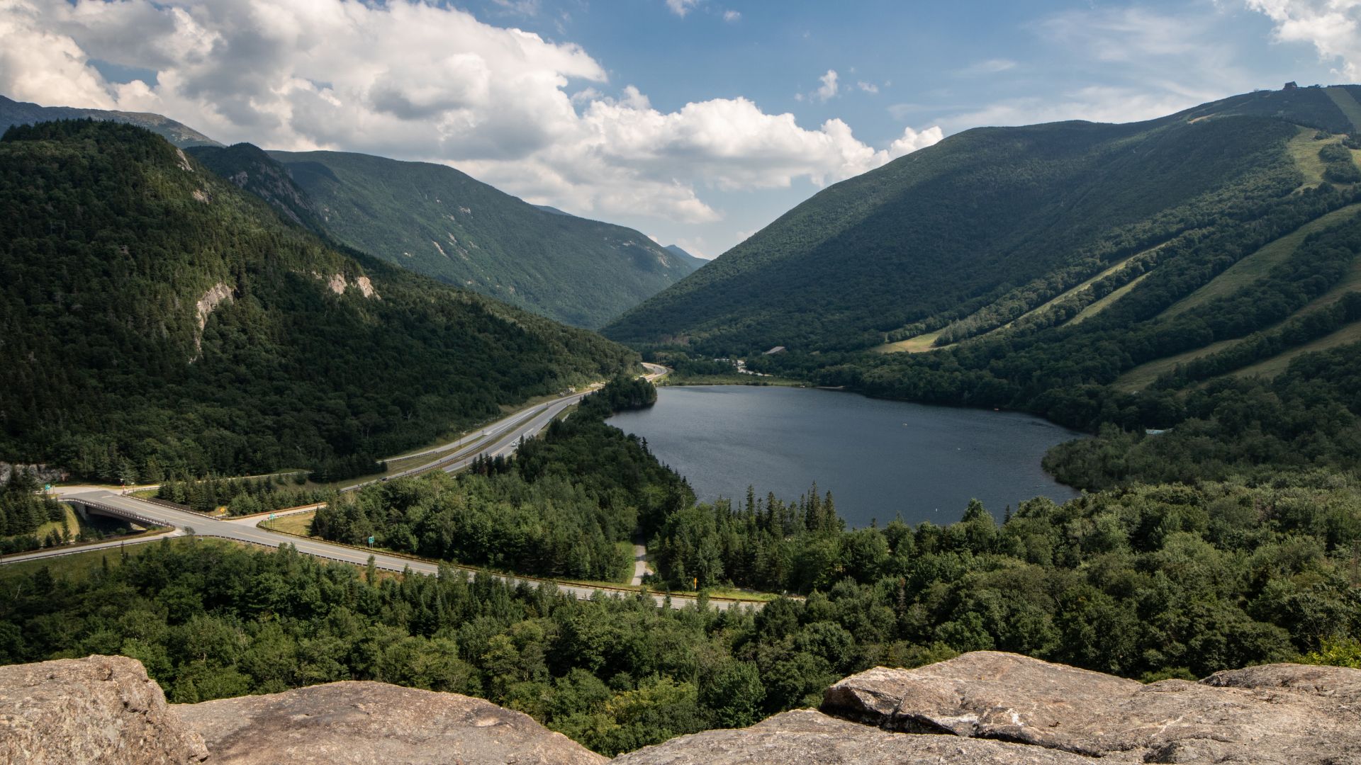 A panoramic view from a rocky cliff overlooking a serene lake and a highway running through a lush, green mountain valley under a partly cloudy sky.