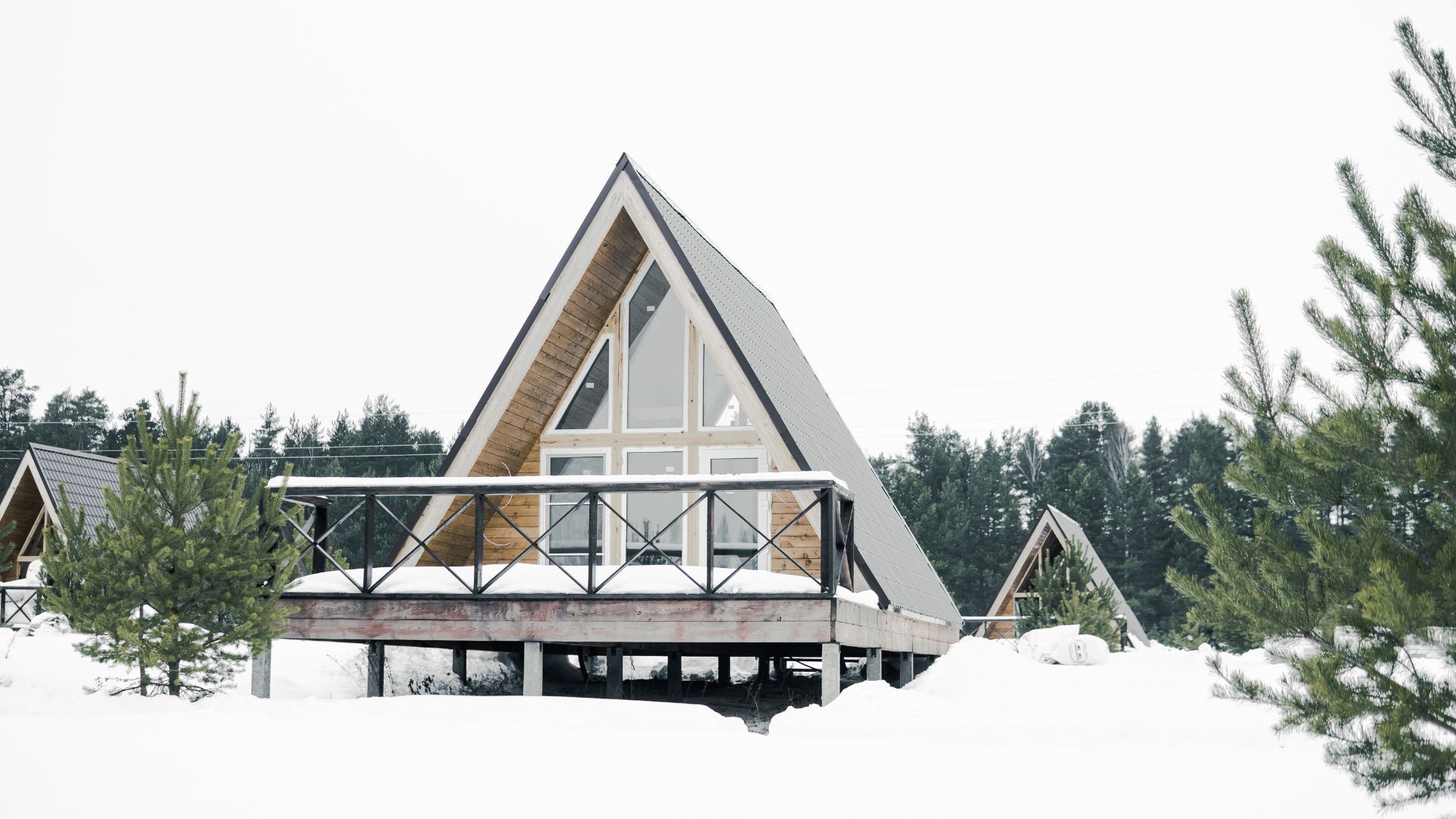 A photograph of a modern wooden A-frame cabin with large windows and a deck, surrounded by deep snow and pine trees.
