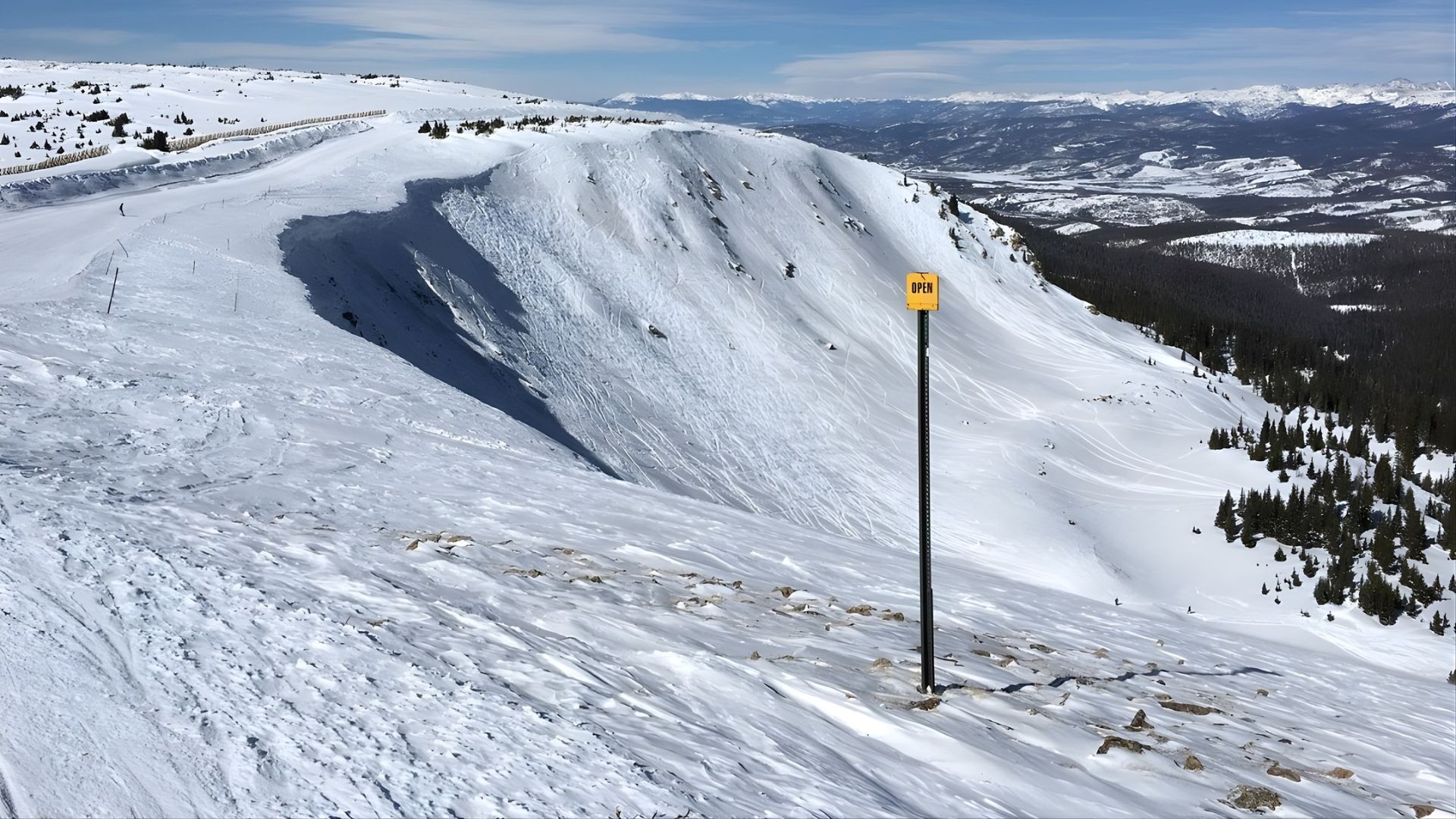 An expansive, elevated view of steep, deep snow-covered mountain slopes under a bright blue sky, with a yellow "OPEN" sign on a tall post in the middle right foreground, and vast mountain ranges visible in the distance.