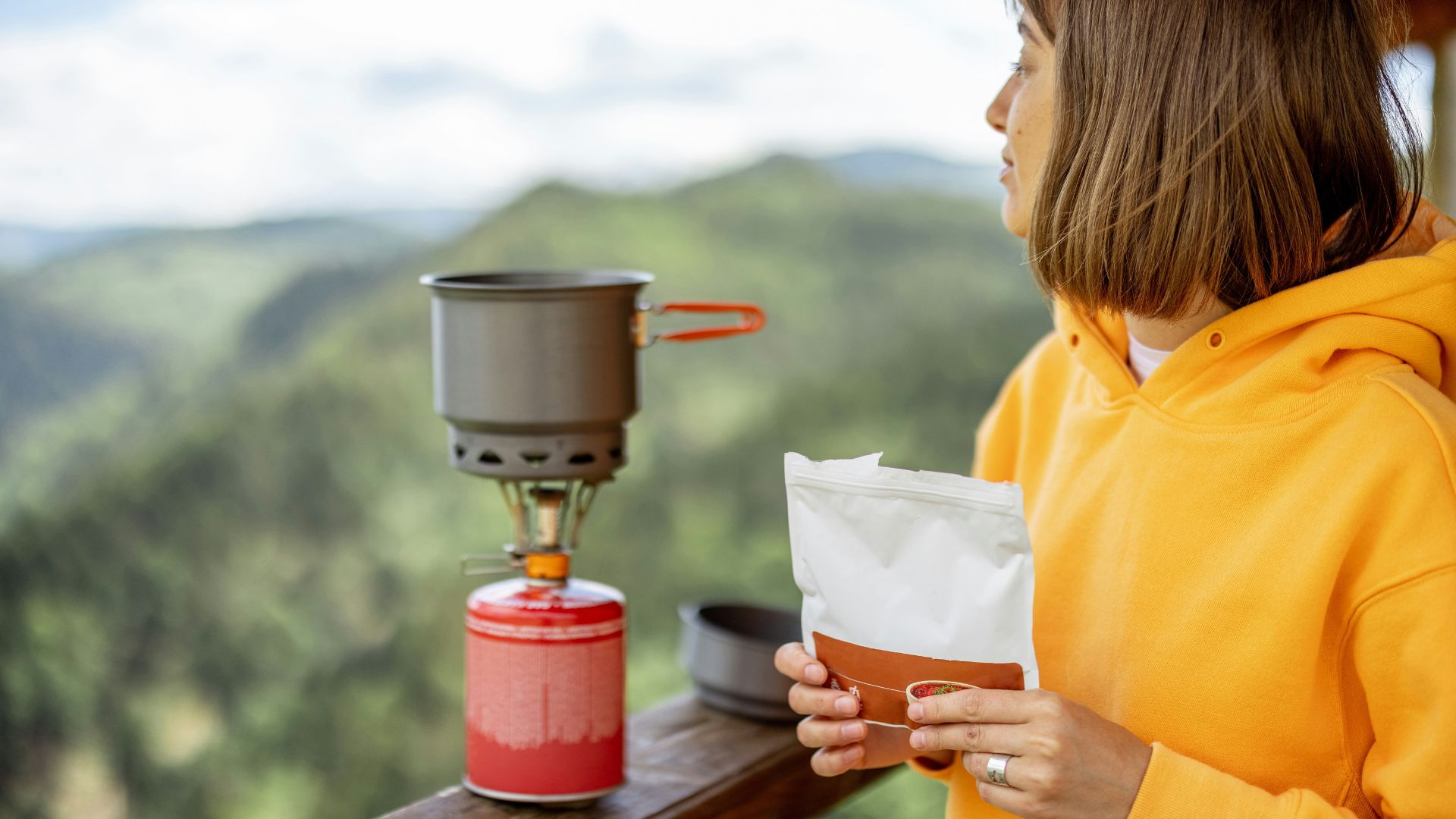 A woman in a yellow hoodie holds a packet next to a camping stove with a pot, overlooking a green, mountainous landscape.