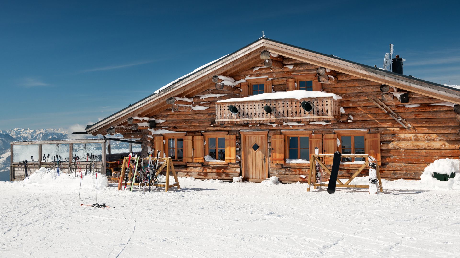 A traditional wooden ski chalet covered in snow with skis and snowboards leaning outside on a sunny day.