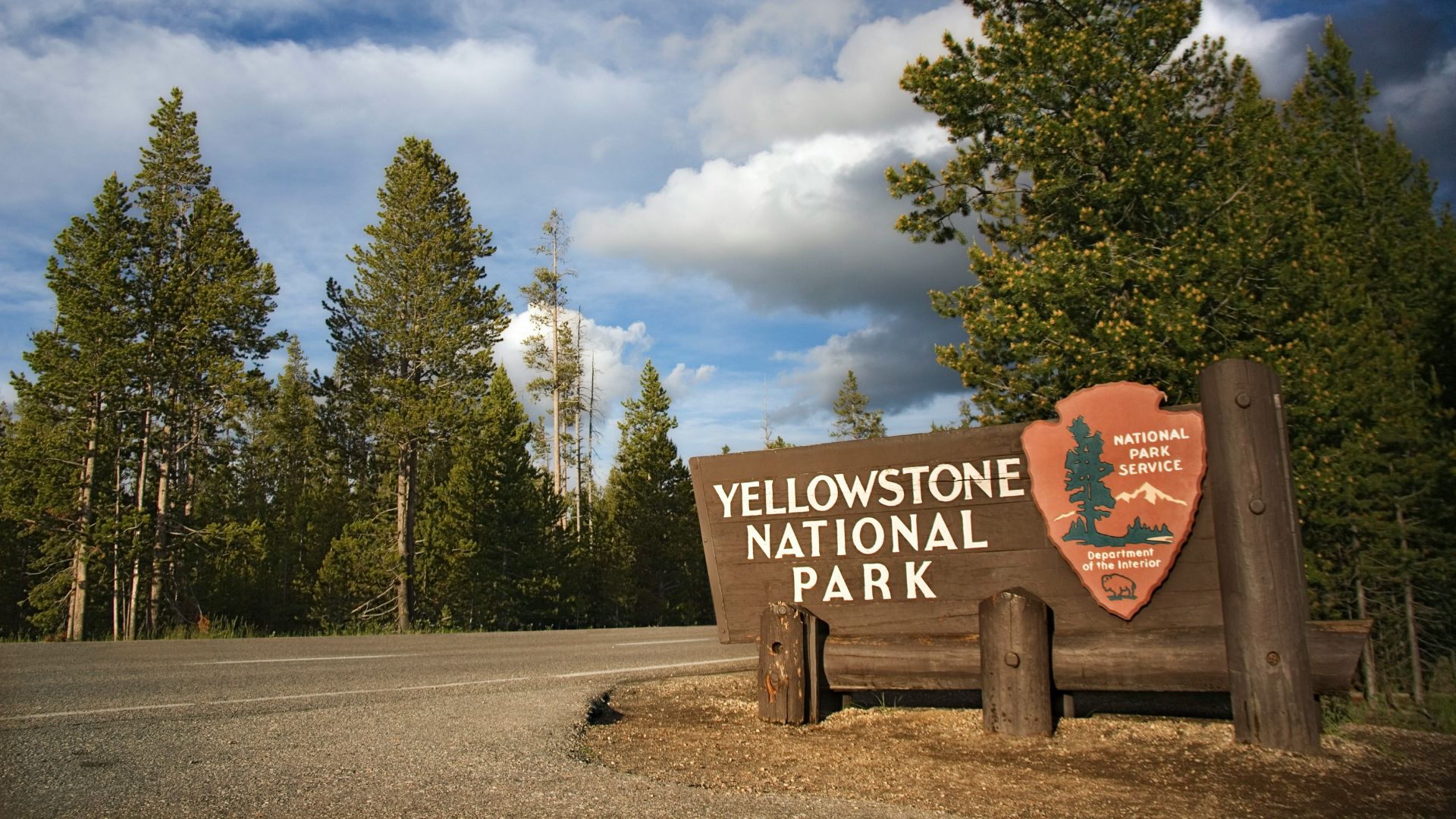 A wide wooden entrance sign for Yellowstone National Park with the National Park Service logo stands next to a paved road under a blue sky with white clouds, surrounded by dense evergreen trees.