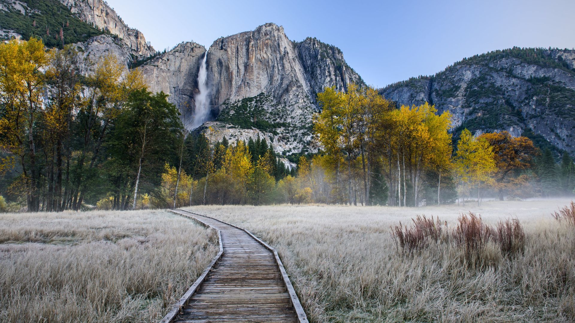 A wooden boardwalk leads through a frosty meadow with yellow and green autumn trees towards a large waterfall (Yosemite Falls) cascading down a massive granite cliff face.