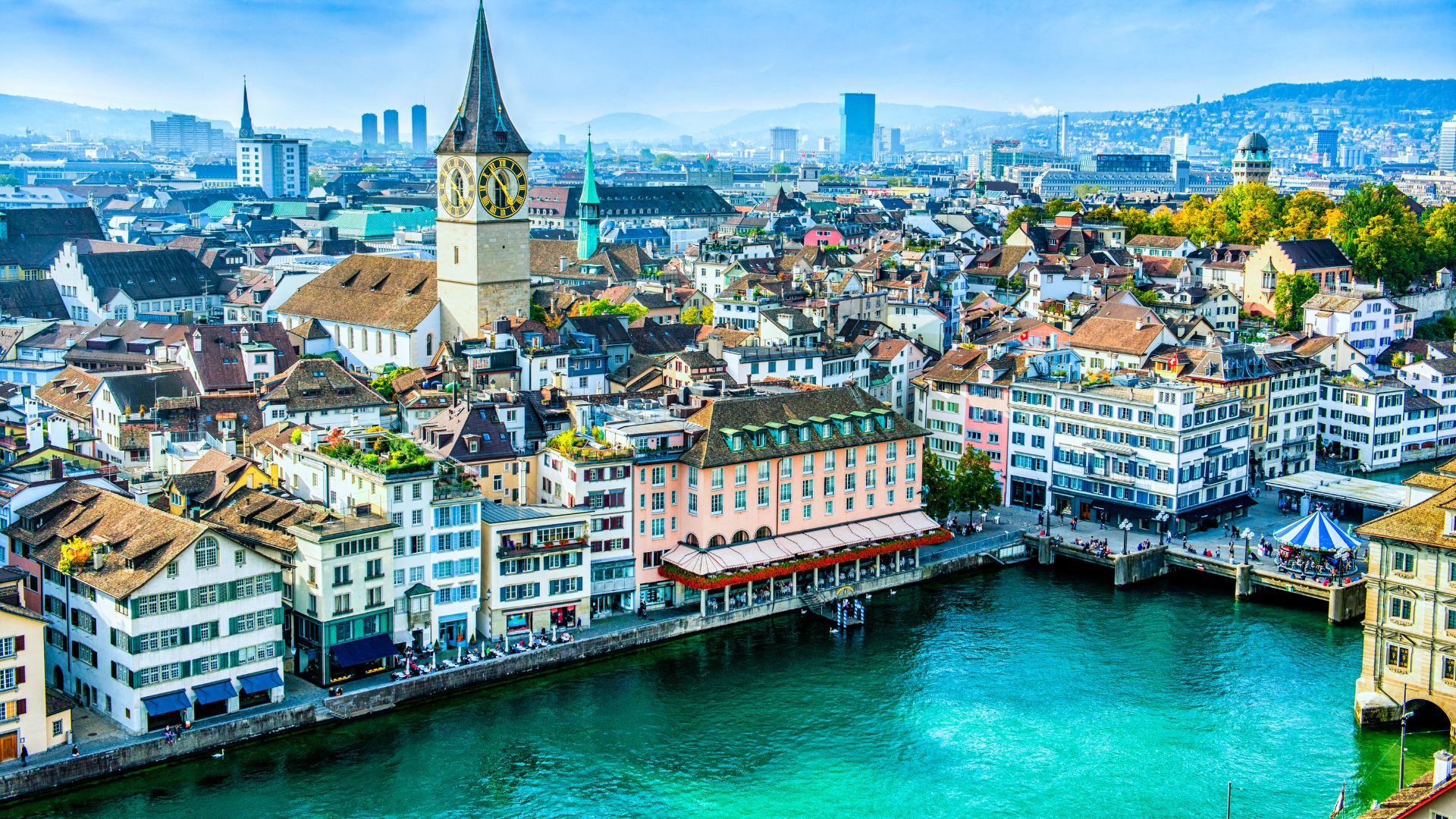 An elevated, panoramic photograph of the historic city of Zurich, Switzerland, showing colorful buildings lining the banks of the Limmat River and the iconic St. Peter's Church clock tower rising above the rooftops.