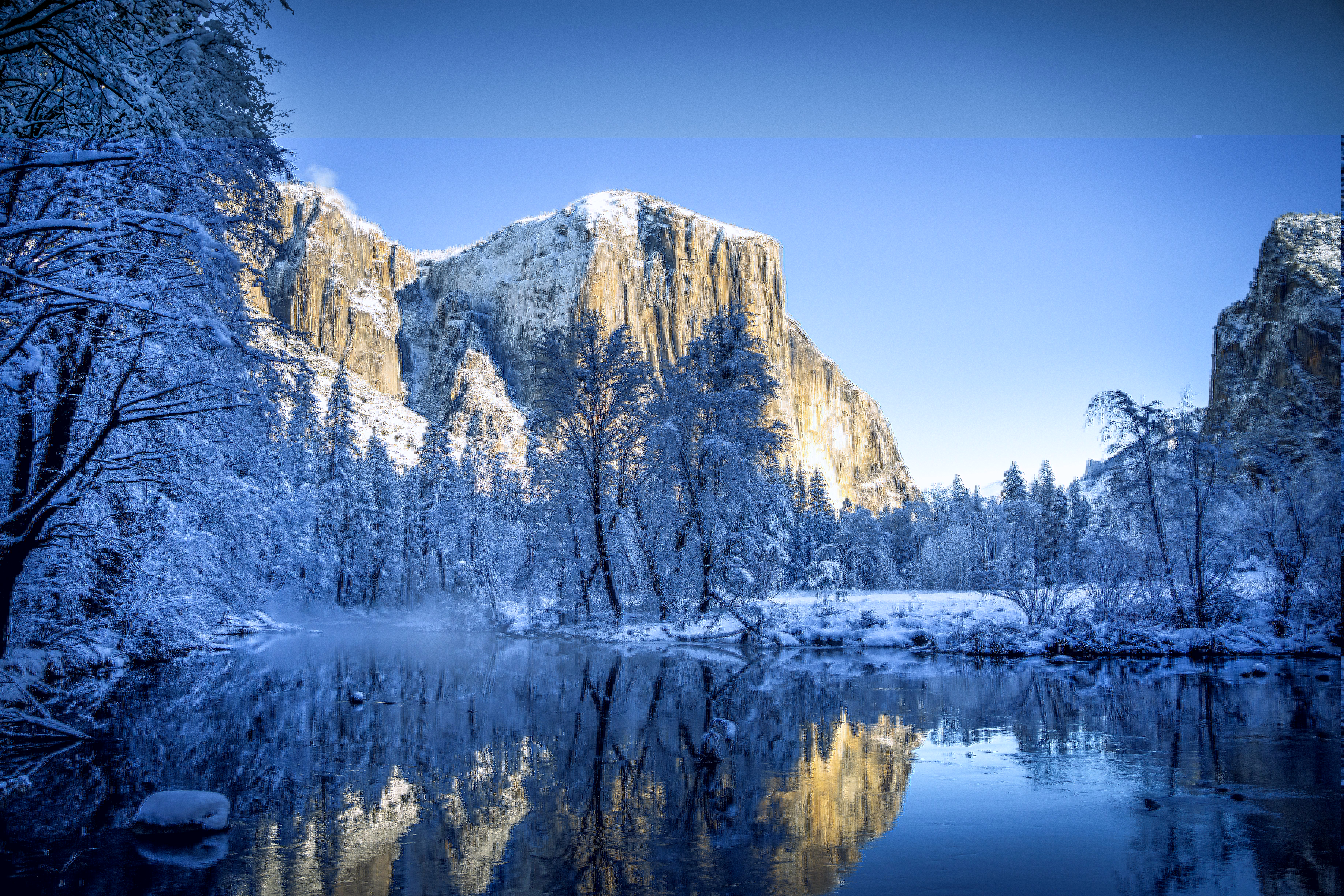 A panoramic view of the snow-covered Yosemite Valley in winter, showing the majestic El Capitan mountain reflected in the calm waters of the Merced River.